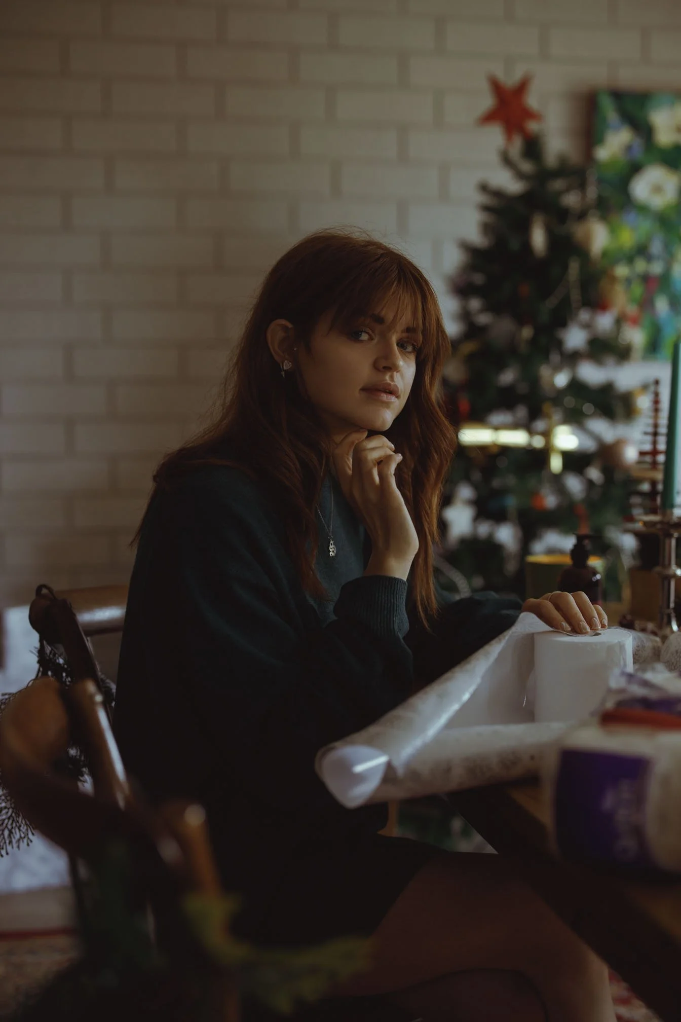 A woman with red hair sitting at a table in front of a decorated Christmas tree, looking at the camera, with holiday decorations in the background.