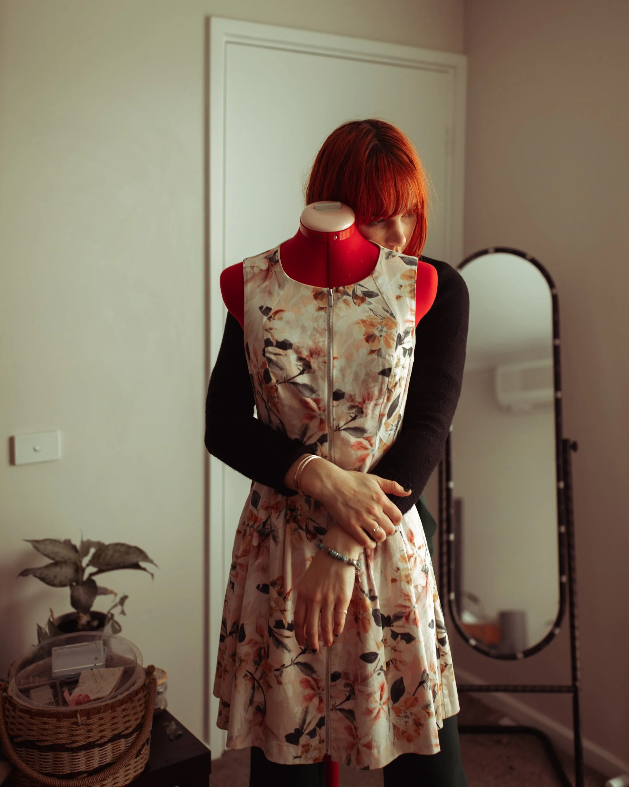 A woman with red hair, wearing a floral dress and black sleeves, stands with her arms crossed in front of a dress form with a floral top. She is inside a room with a mirror and a plant on a table.