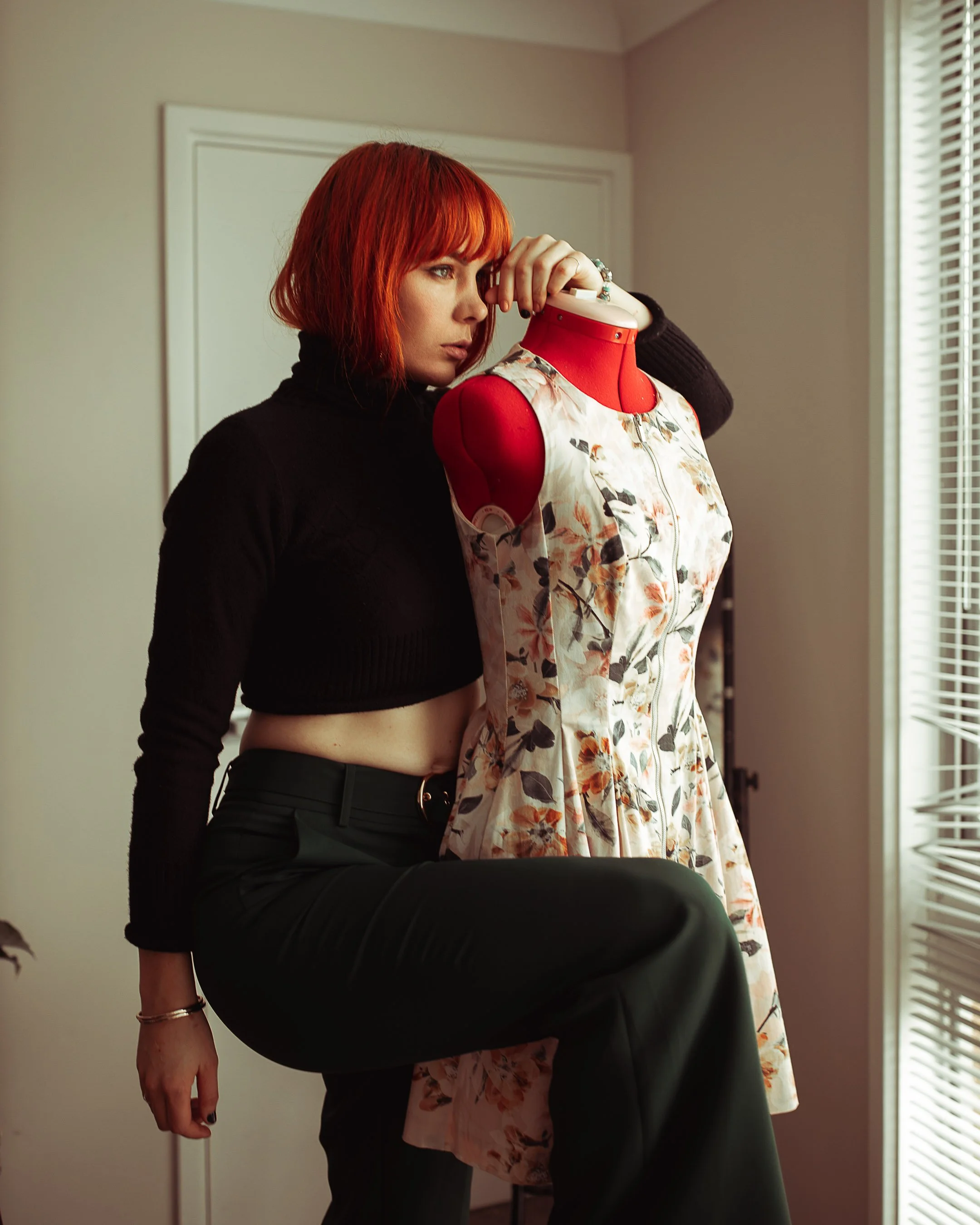A woman with red hair and a black crop top sitting on a table, leaning against a mannequin dressed in a floral dress, near a window with blinds.