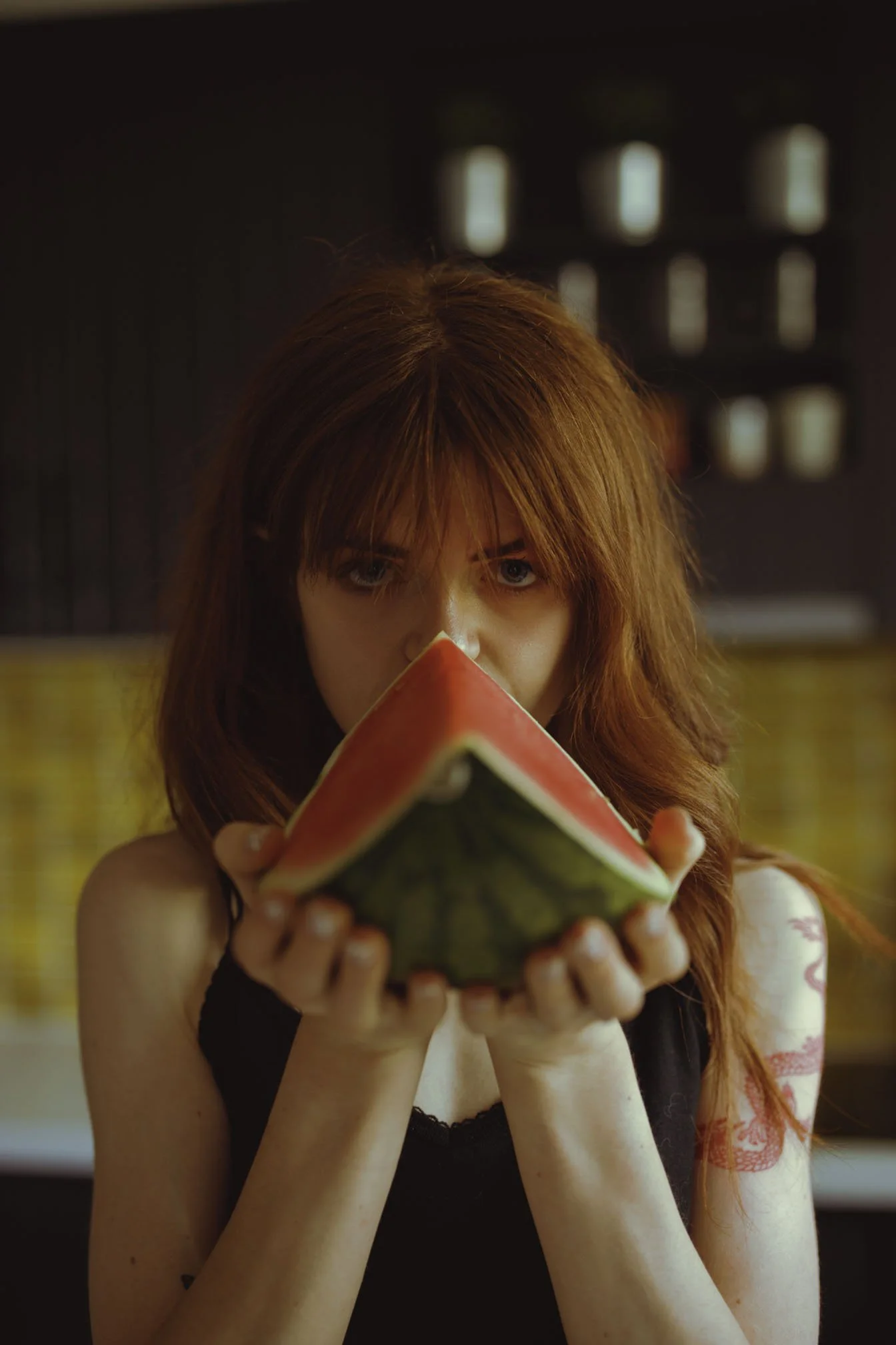 A woman with red hair holds a slice of watermelon close to the camera, looking directly at the viewer.