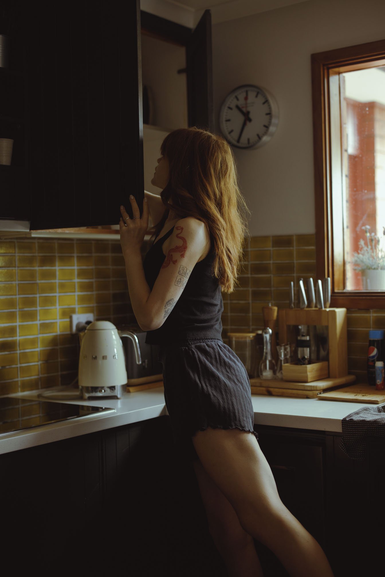 A woman with long red hair and tattoos on her arm, wearing a black tank top and shorts, standing in a kitchen, opening a cabinet.