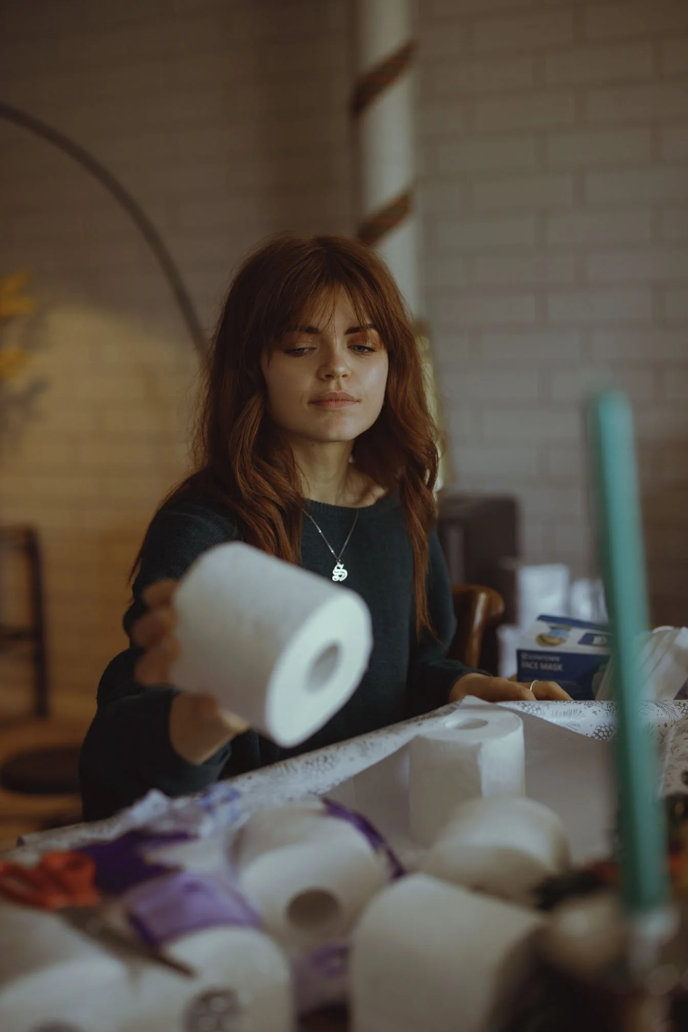 A young woman with red hair and a black sweater is sitting at a table, holding a roll of toilet paper in her right hand. There are multiple rolls of toilet paper on the table in front of her. The background includes a brick wall and some household it