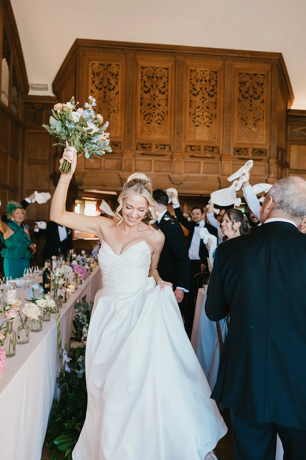 A bride in a white wedding dress smiling and celebrating with her bouquet raised, surrounded by guests at a wedding reception in a wood-paneled room.