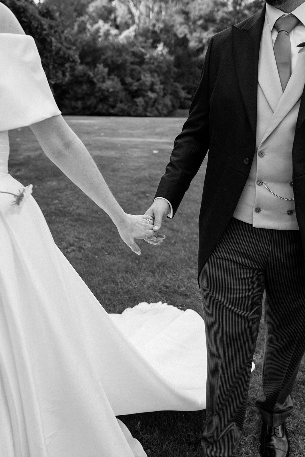A black and white photo of a bride and groom holding hands outdoors, with the bride's wedding gown and the groom's suit visible.