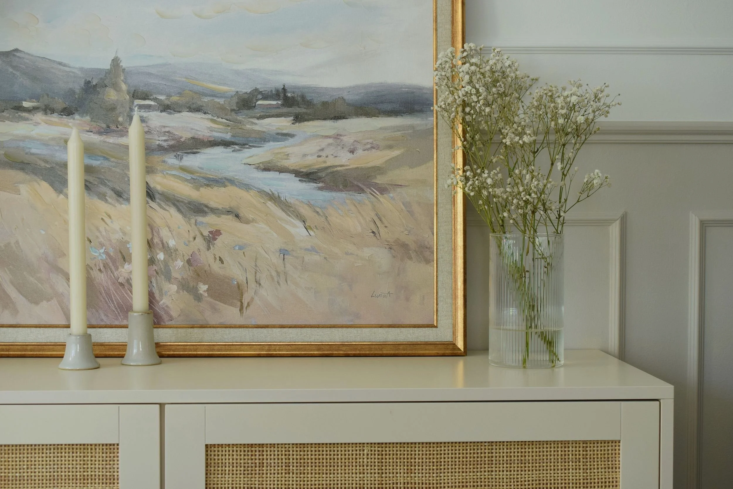A white sideboard with two white candles in vases on it, a framed painting of a landscape, and a clear vase with white baby's breath flowers, all against a light-colored wall.