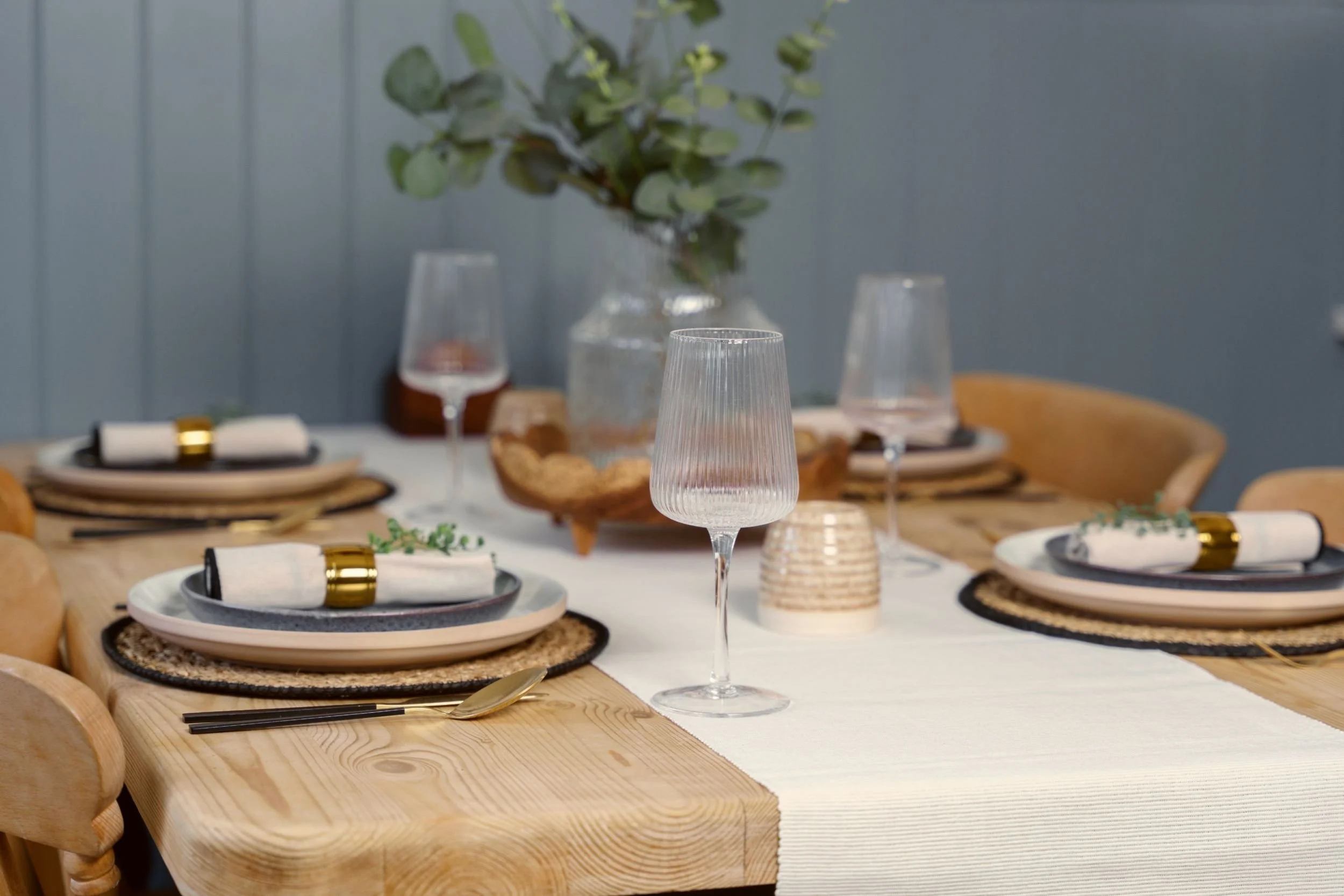 A decorated dining table set with plates, napkins, gold utensils, wine glasses, and a centerpiece with green foliage in a glass vase, with a light-colored tablecloth and wooden chairs surrounding it.