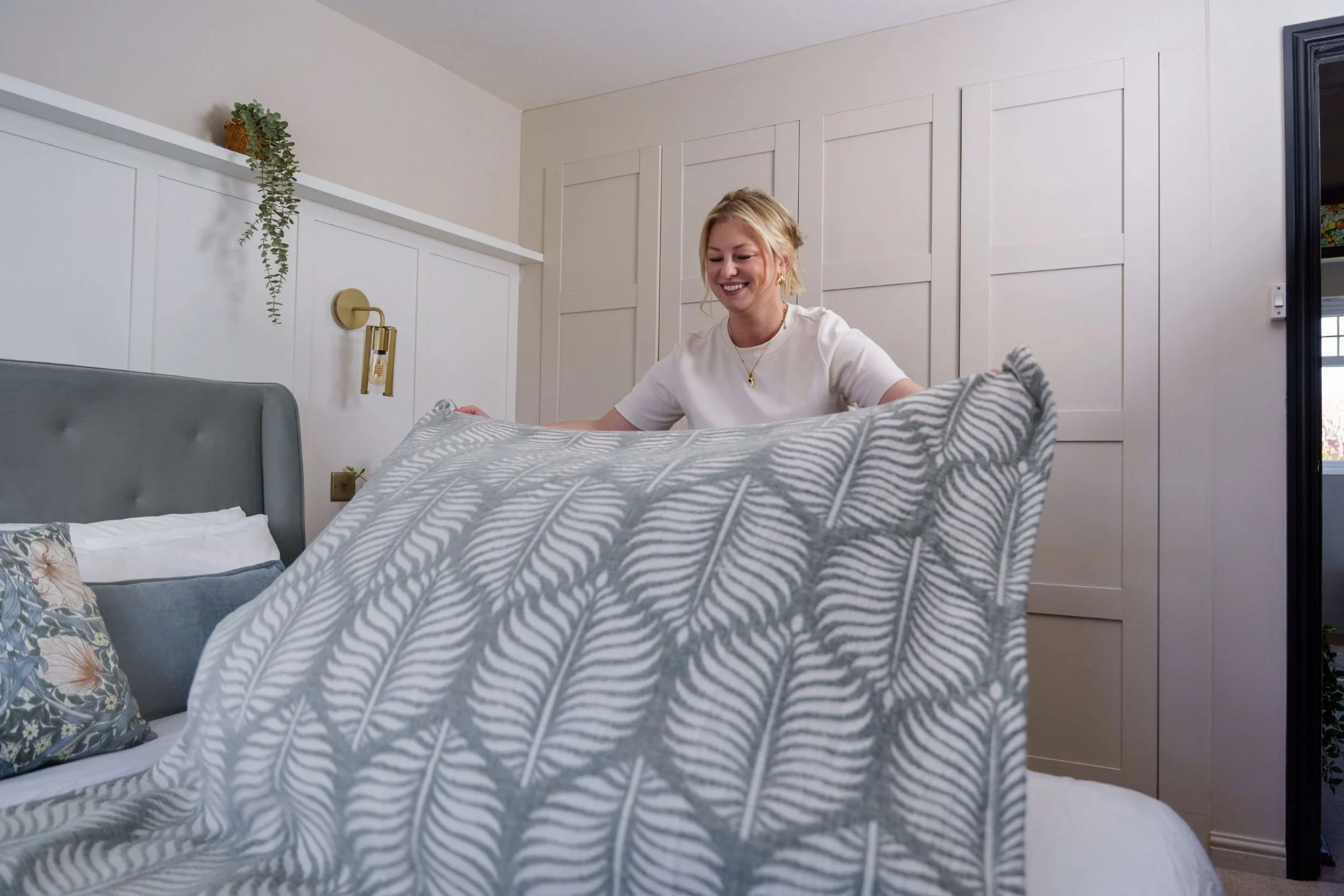 A woman with blonde hair sits on a bed, smiling, in a bedroom with white walls and paneling, holding a pillow with a gray and white leaf pattern.
