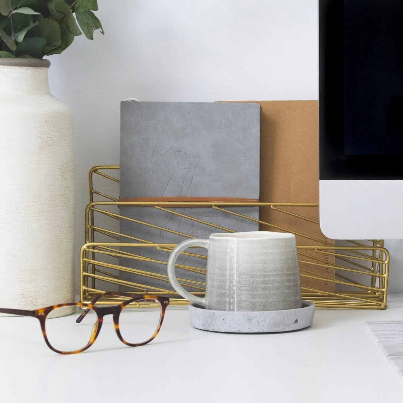 A desk workspace with a white mug, a pair of tortoiseshell glasses, a gold wire desk organizer holding notebooks, a gray notebook, a brown notebook, and a laptop. A green plant in a white textured pot is on the left.