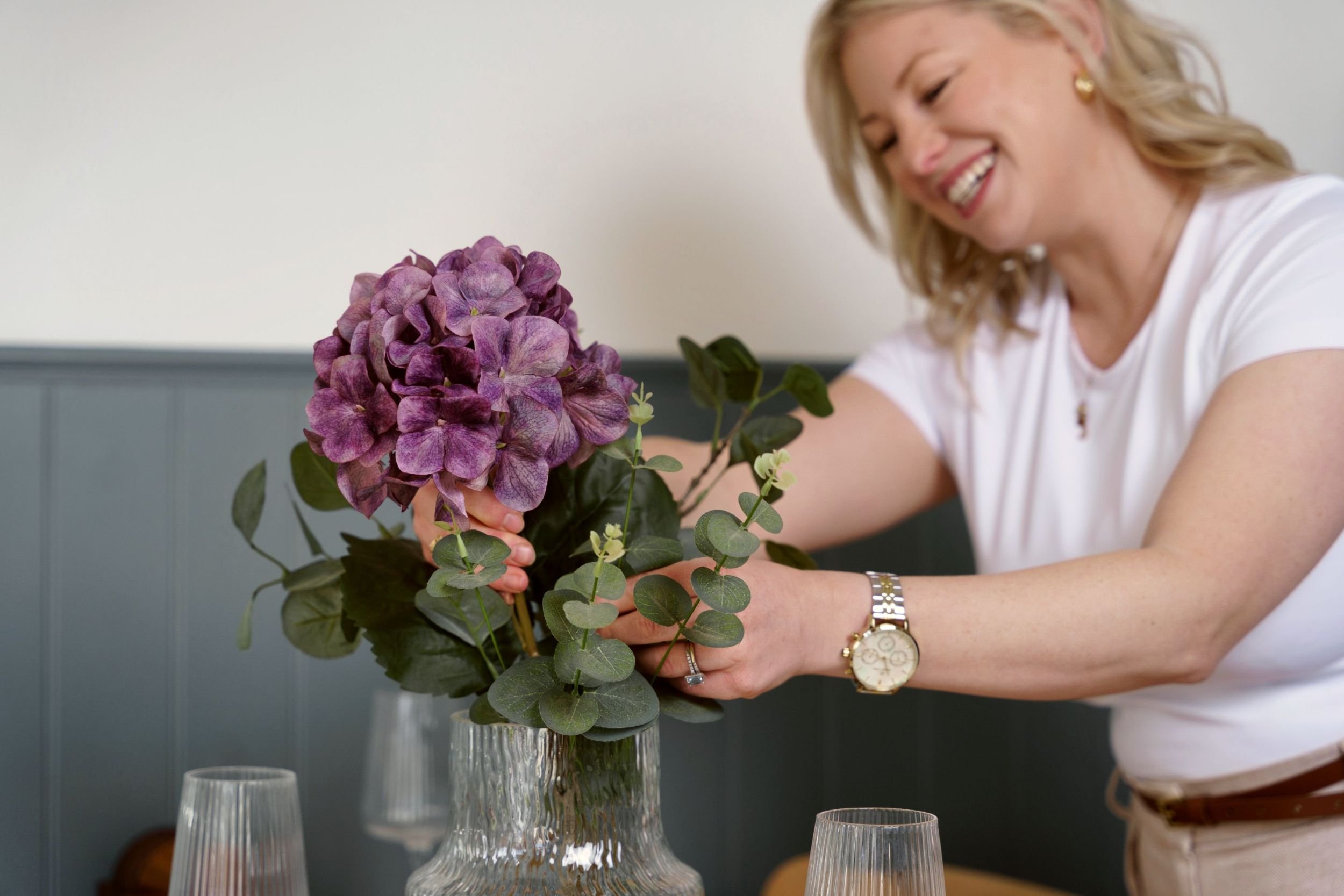 A woman with blonde hair, wearing a white shirt and gold earrings, arranging a bouquet of purple hydrangeas and green foliage in a silver vase at a dining table.