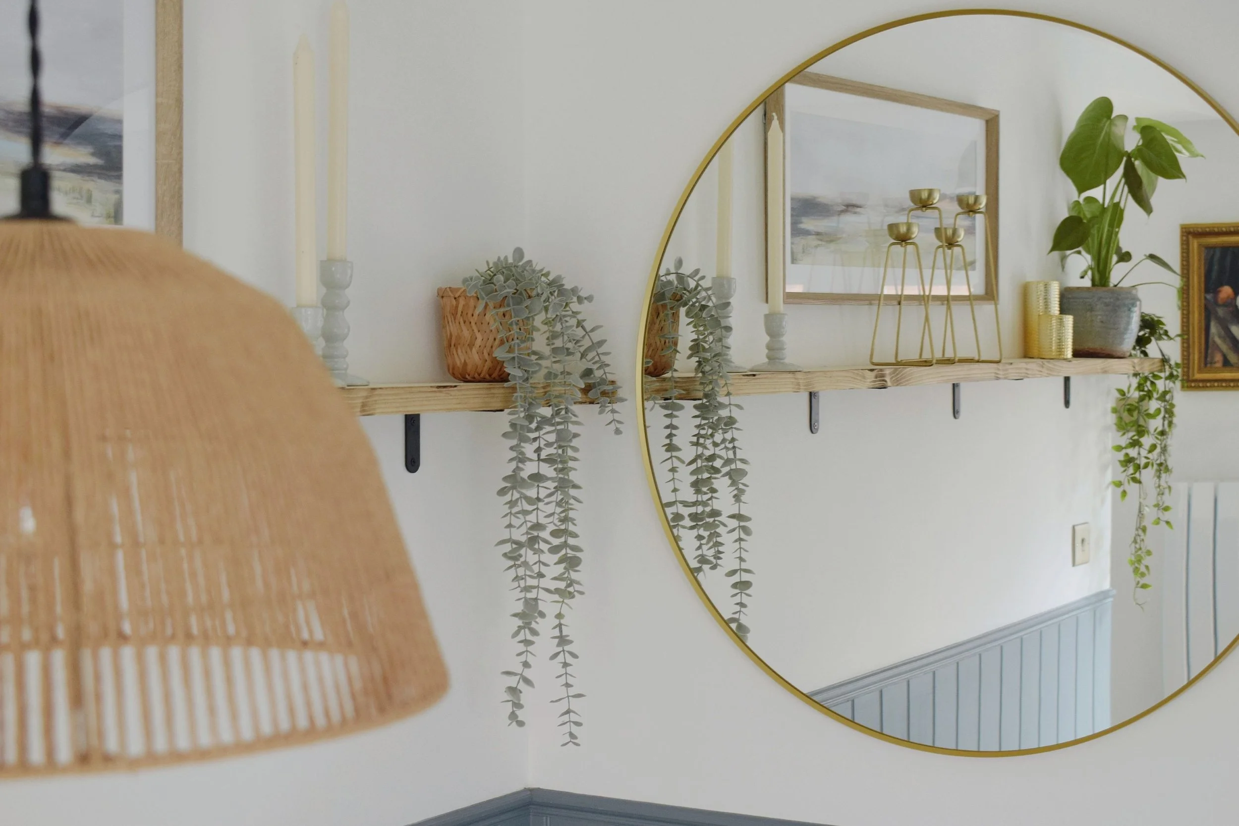 Decorative wall shelf displaying candles, plants, and artwork, with a large round mirror reflecting part of the shelf and artwork, in a modern interior setting.