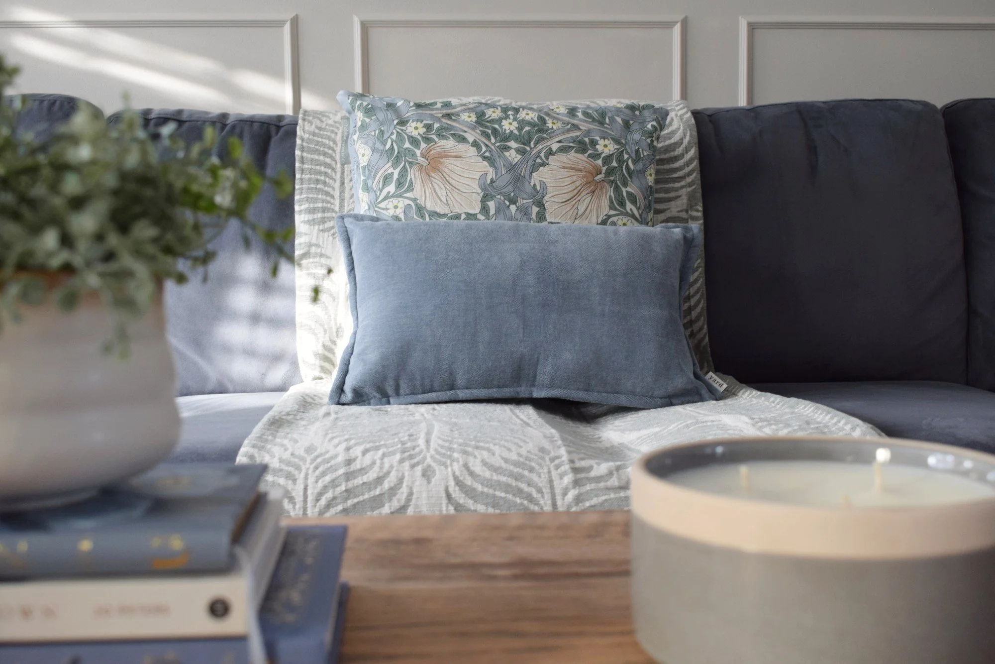 A cozy living room sofa with multiple pillows, including a floral-patterned pillow, a light blue pillow, and a dark blue pillow, under a white paneled wall. A flower arrangement in a white pot is partially visible in the foreground, and a tray with a mug and coasters is on a wooden table nearby.