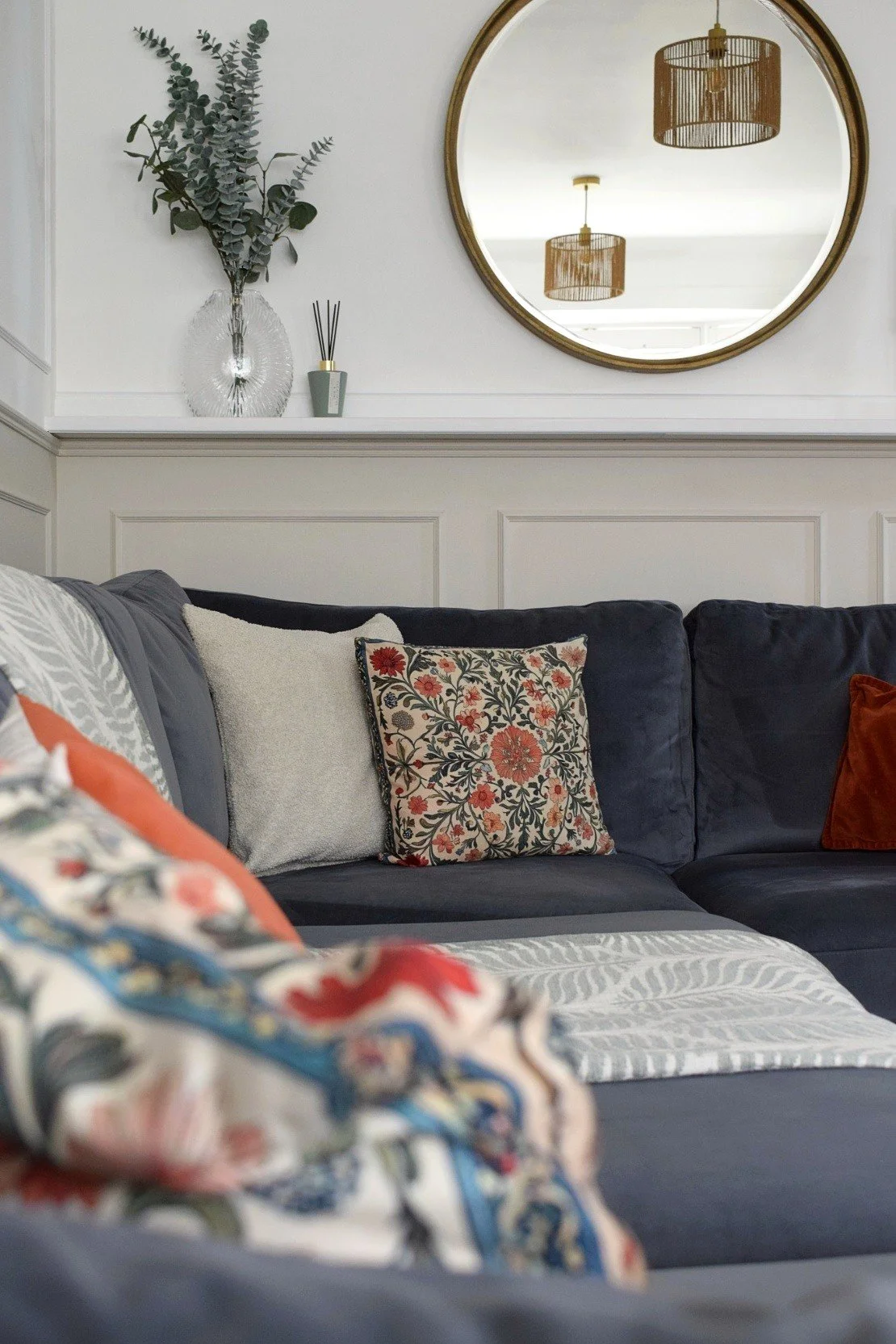 A living room corner with a navy blue sofa, decorative pillows, a wall mirror, wall baseboard and a vase with green eucalyptus leaves. A ceiling light fixture is reflected in the mirror.