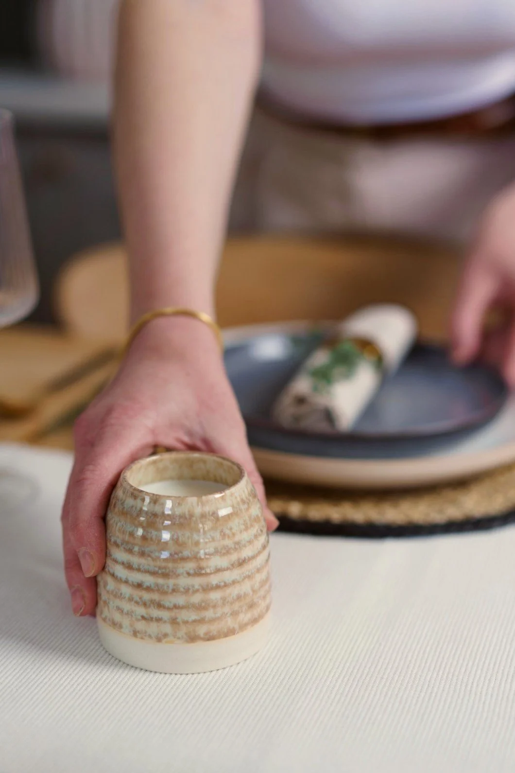 Person holding a ceramic cup with beige and white stripes, filled with a beverage, with a table set of dishes and a wrapped food item in the background.