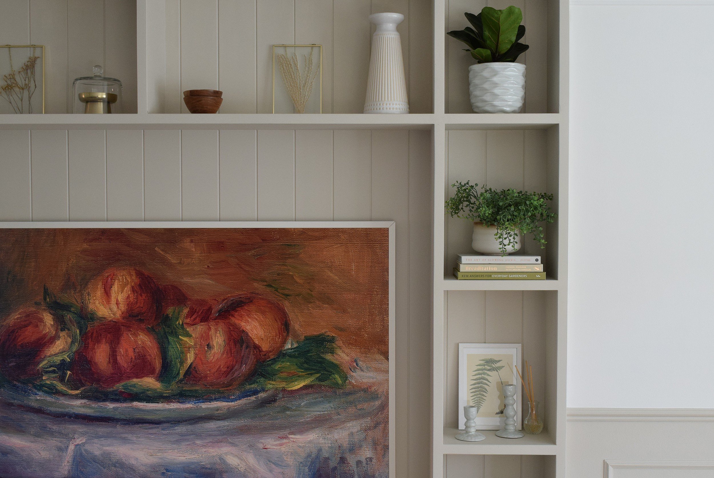A white shelving unit with decorative items including a glass jar with a lid, a small brown bowl, a framed dried plant, a tall white vase, a potted plant, a stack of books, and framed artwork, with part of a colorful oil painting of roses visible at the bottom.