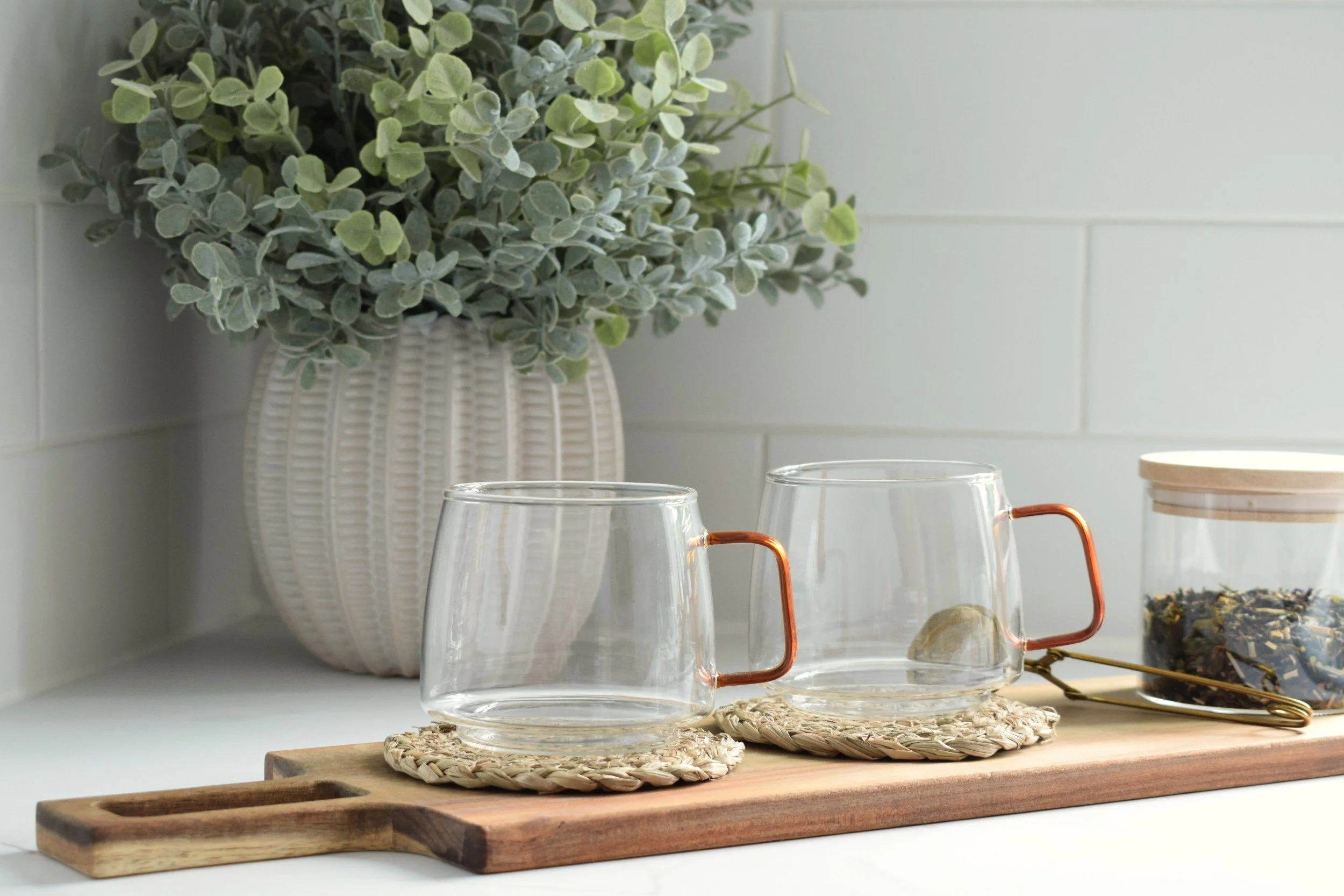 A white ceramic vase with a textured surface holds a green leafy plant. In front of the vase are two clear glass cups with copper handles, placed on woven round coasters on a wooden tray. A wicker woven mat and a pair of black sunglasses with gold accents are on the tray. The background consists of a white tiled wall.