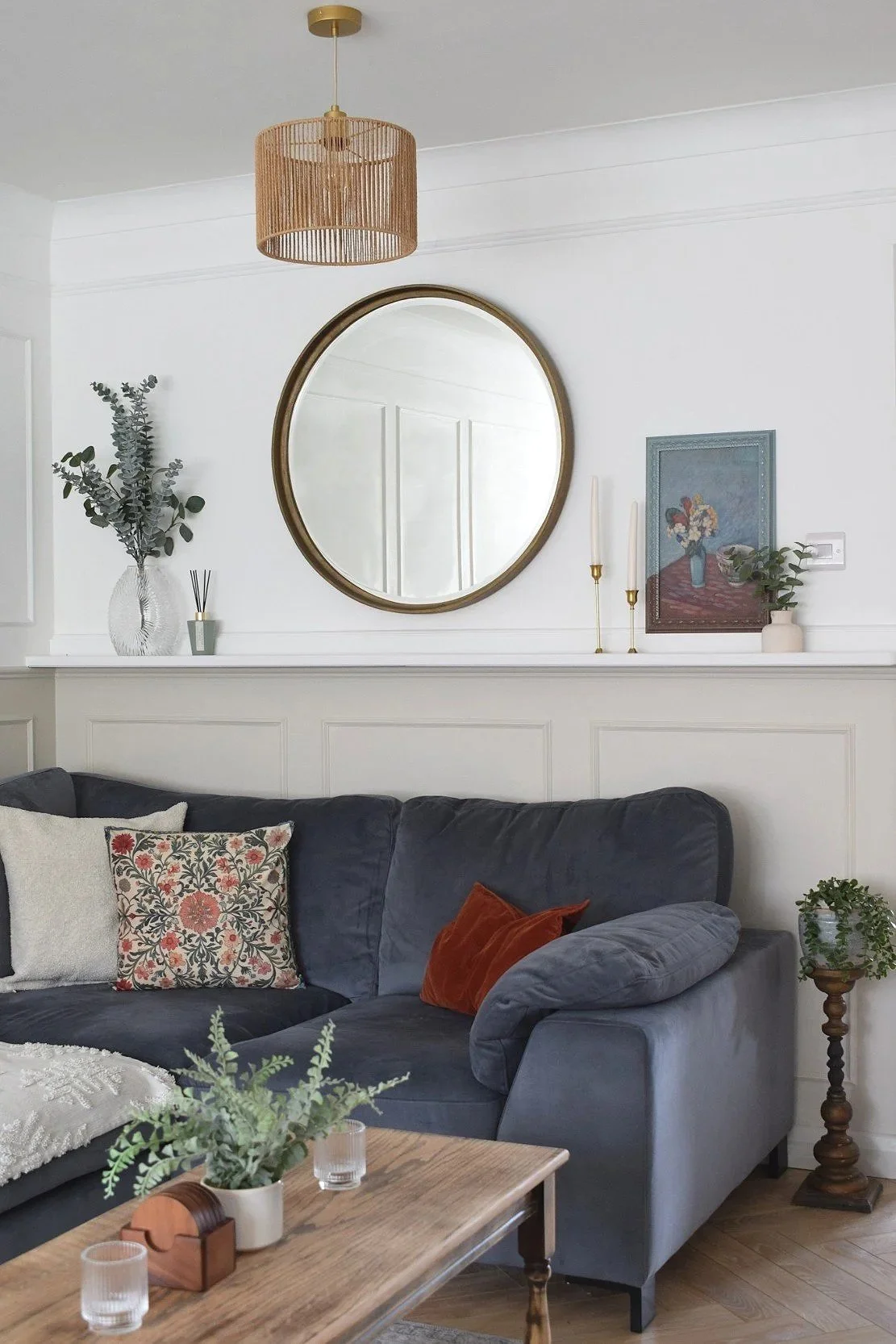 Living room with gray sectional sofa, decorative pillows, wooden coffee table with plants and candles, white wall with a large round mirror, framed art, candles, and vases, and a ceiling light fixture.