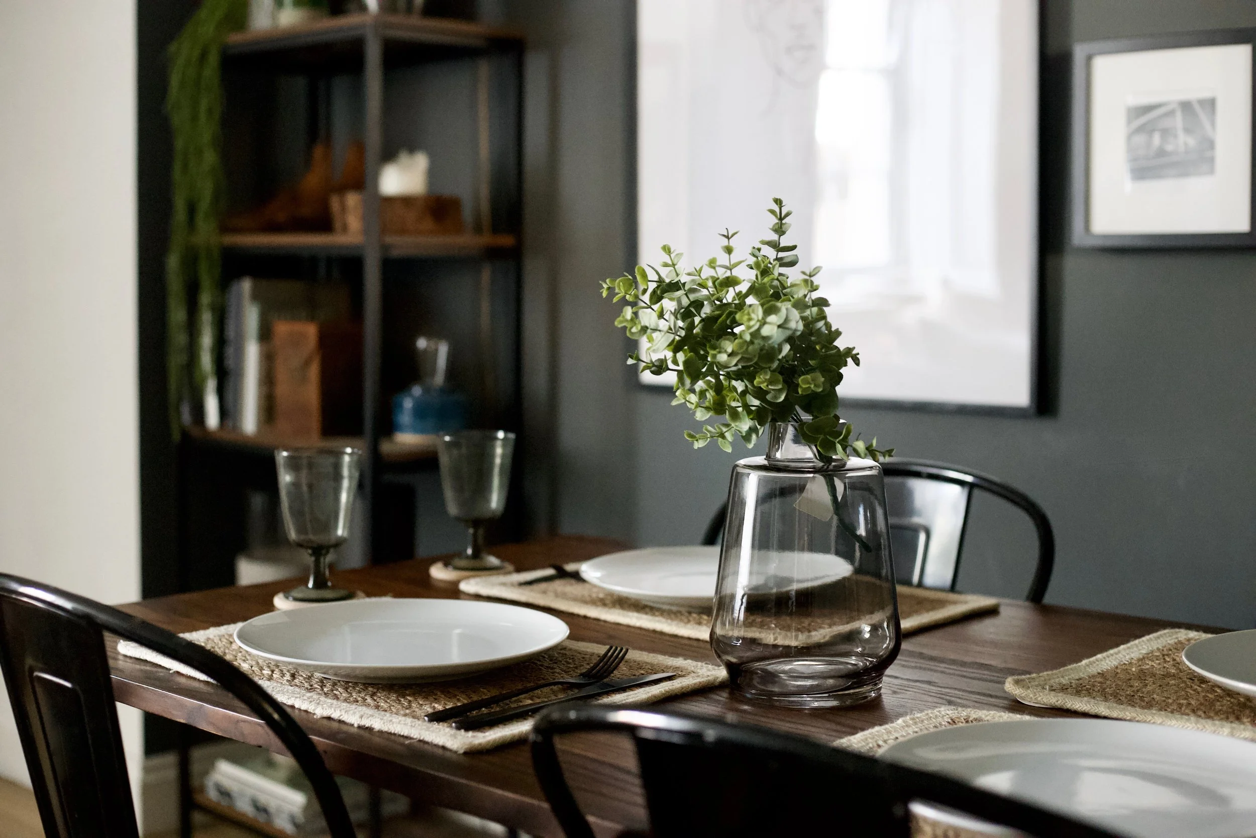 A dining table set for a meal with white plates, silverware, and glassware. A small potted plant is in a glass vase at the center of the table. The background features a dark wall, framed black and white photographs, and a wooden shelving unit with decorative items.