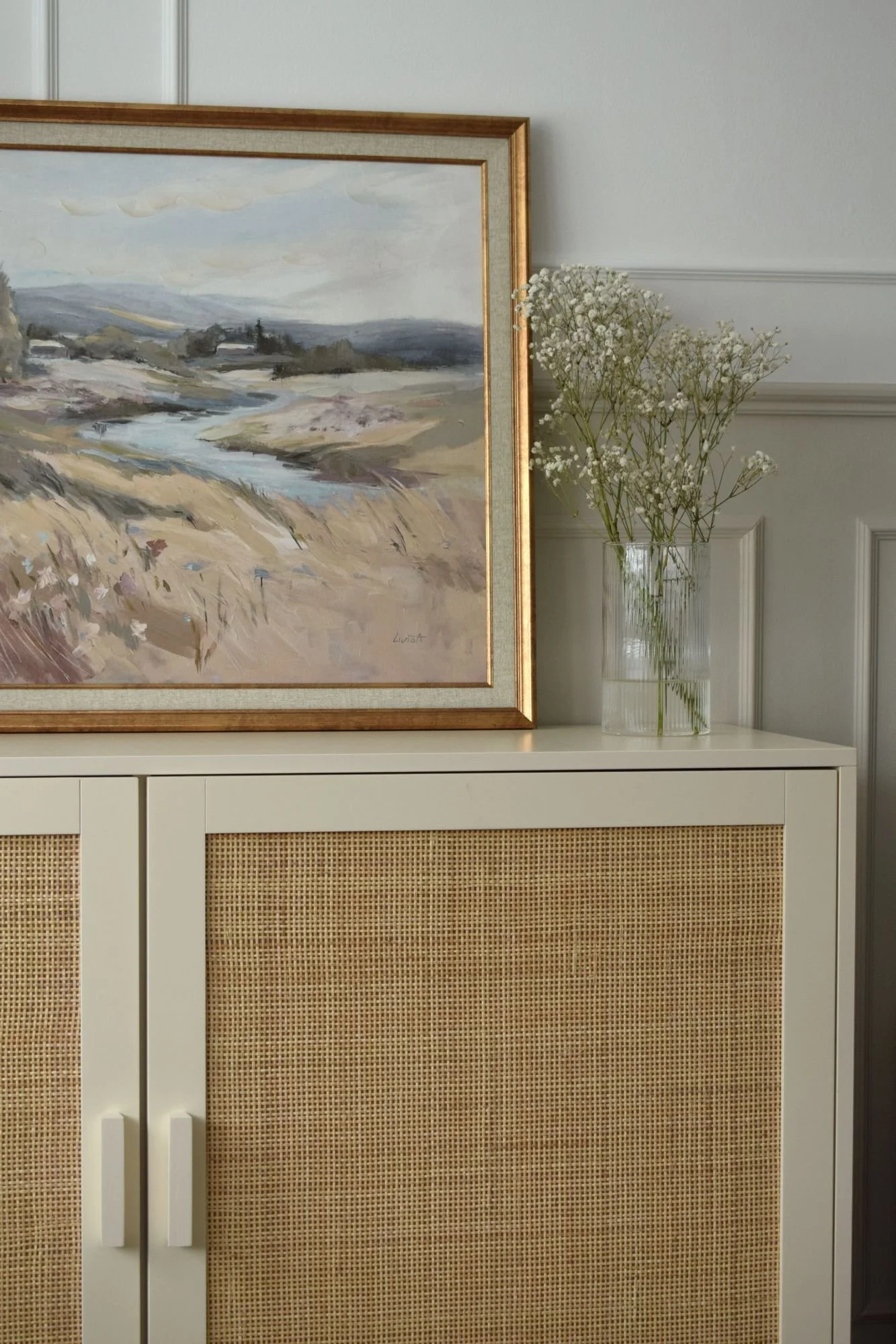 A decorative cabinet with a white top and rattan panel doors, a tall clear glass vase filled with white baby’s breath flowers, a framed watercolor landscape painting of a river and hills in soft pastel tones hanging on the wall.