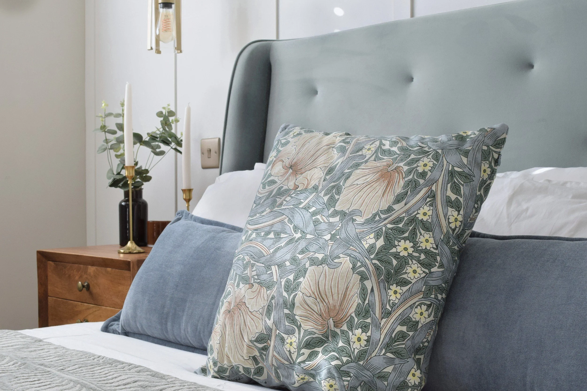 A bedroom with a bed, decorative pillows with floral patterns, a wooden bedside table, a brass candlestick with two white candles, a black vase with greenery, and an IV drip hanging on the wall.