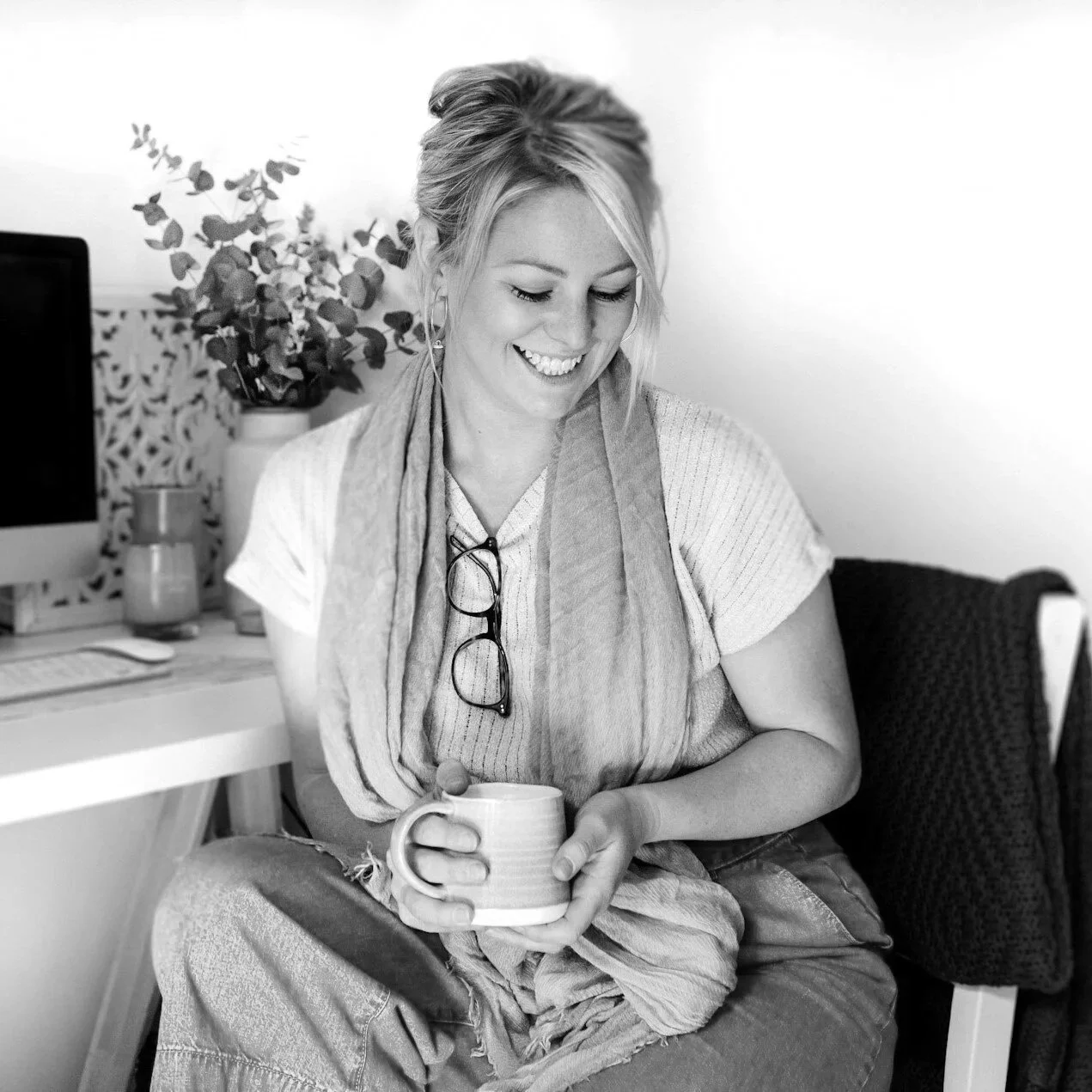 Smiling woman sitting in an office, holding a mug, with glasses hanging from her shirt collar, and plants and computer on the desk behind her.