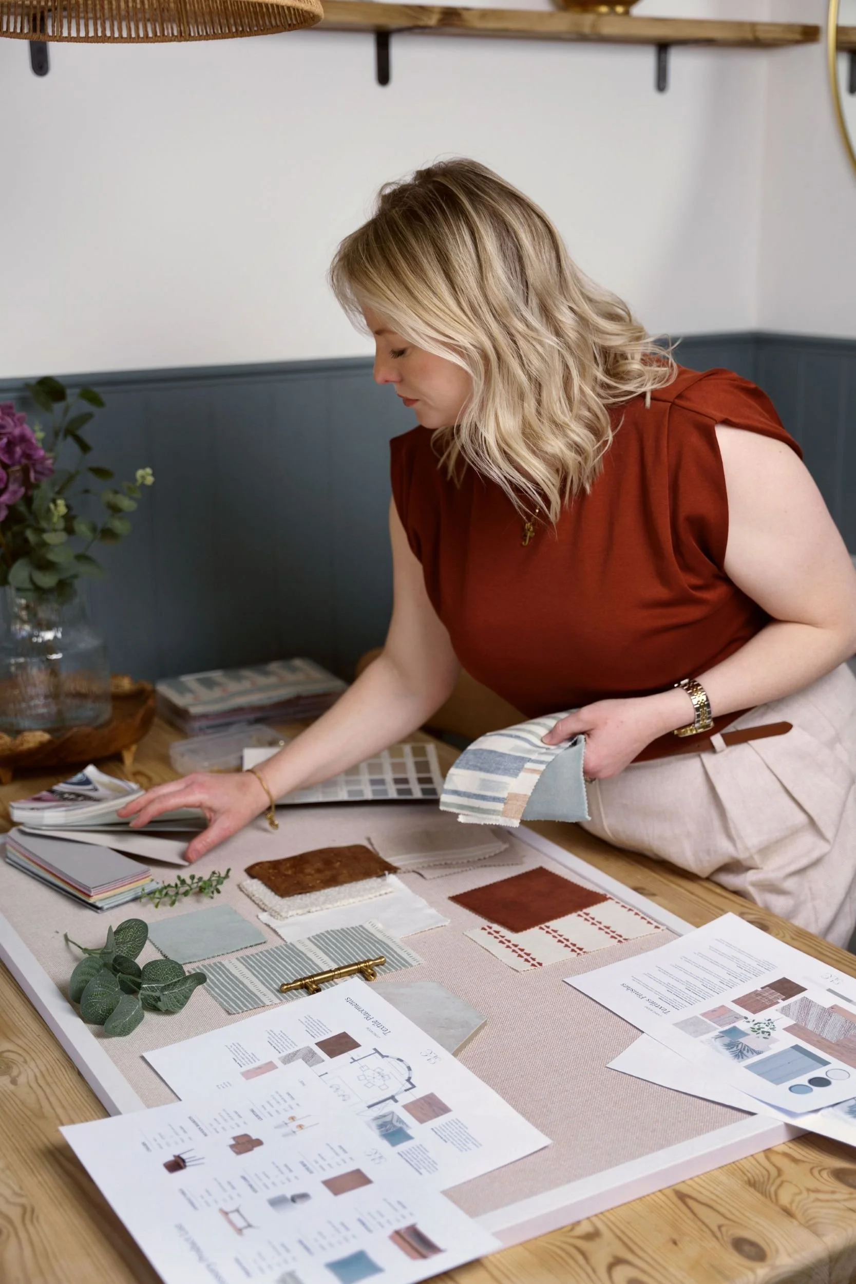 A woman with blonde hair is laying fabric samples and interior design materials on a table, with a flower arrangement and magazines nearby in a well-lit room.