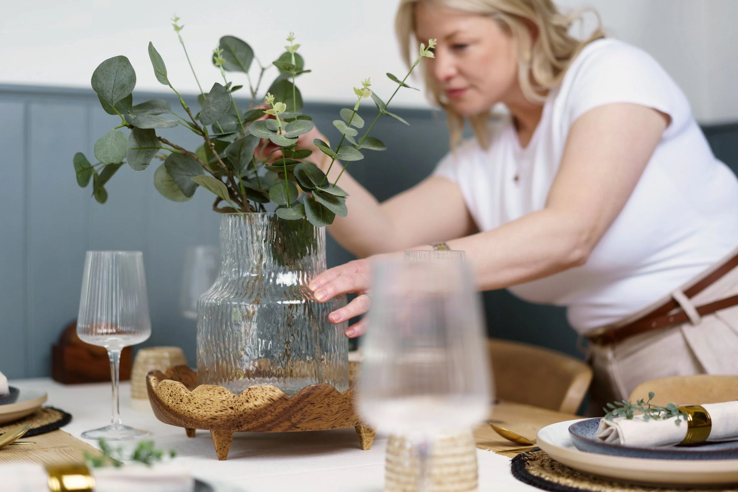 A woman arranging a green leafy plant in a textured glass vase on a dining table set with wine glasses and various dishes.