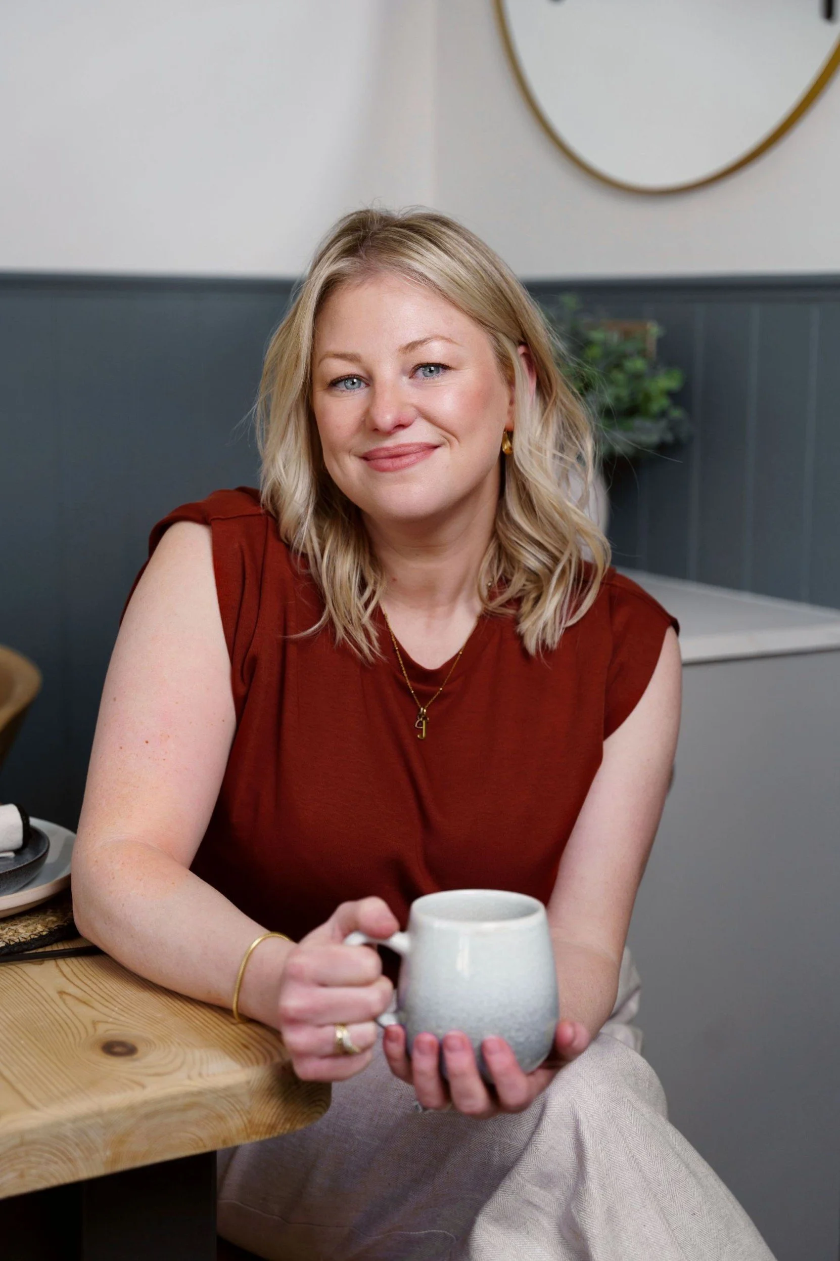 A woman with blonde hair wearing a rust-colored sleeveless top holding a white ceramic mug at a wooden table in a cozy room with a plant and a mirror in the background.