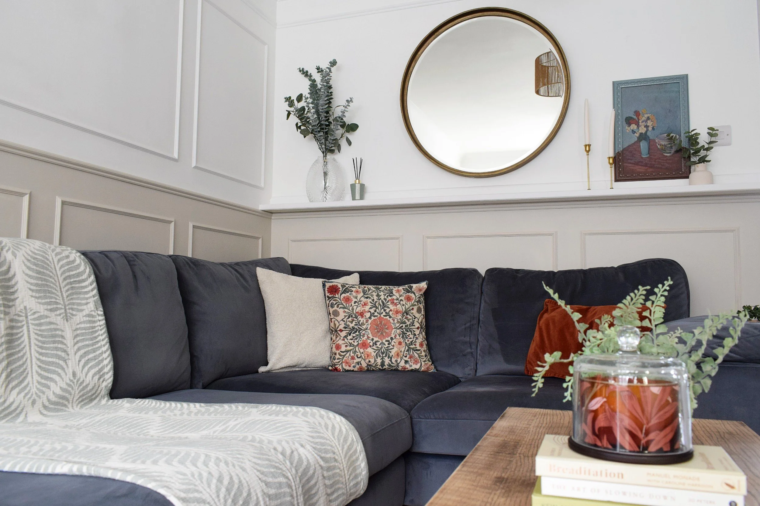 A living room with a dark blue sectional sofa, decorative pillows, a wooden coffee table, a glass jar with a floral candle, wall art, a large round mirror, a vase with eucalyptus, and a framed picture on a white wall.