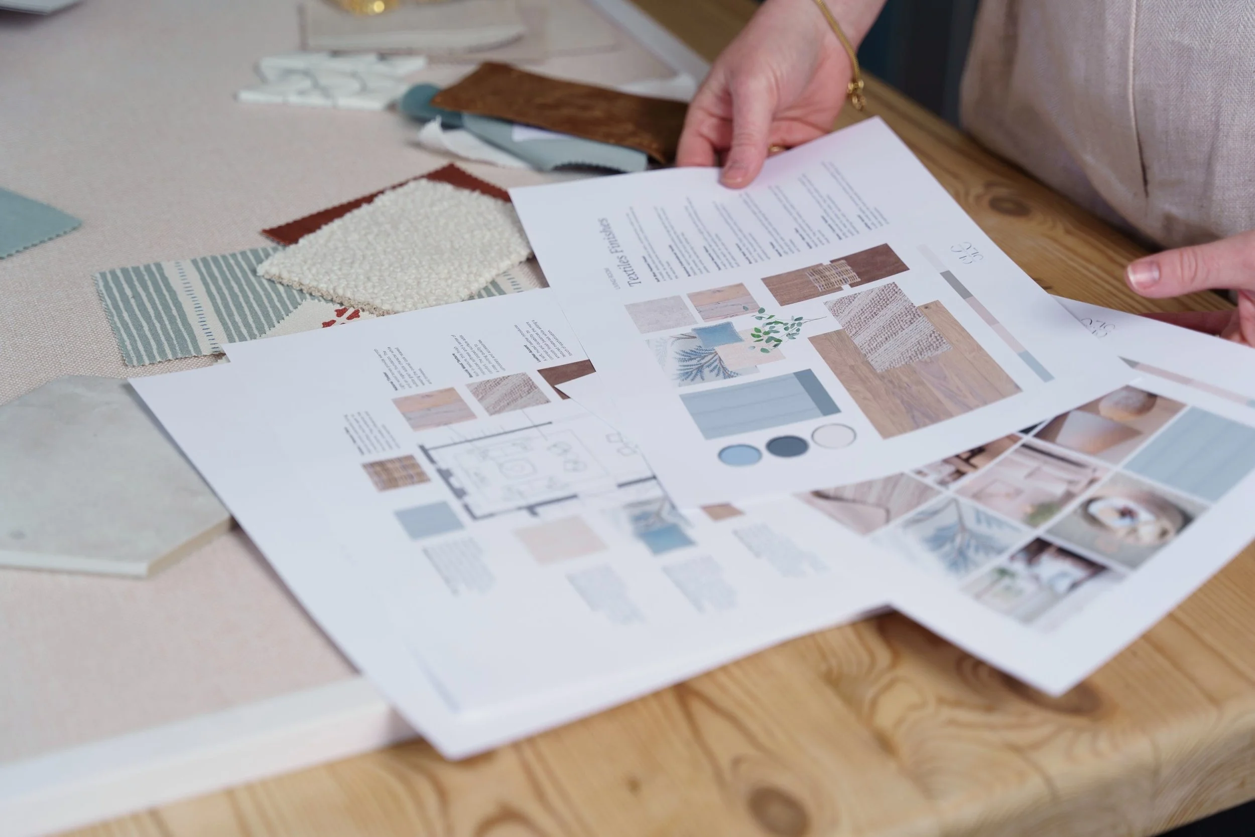 Person examining a color and material palette for home decor, with fabric samples, a catalog, and a tool on a wooden table.