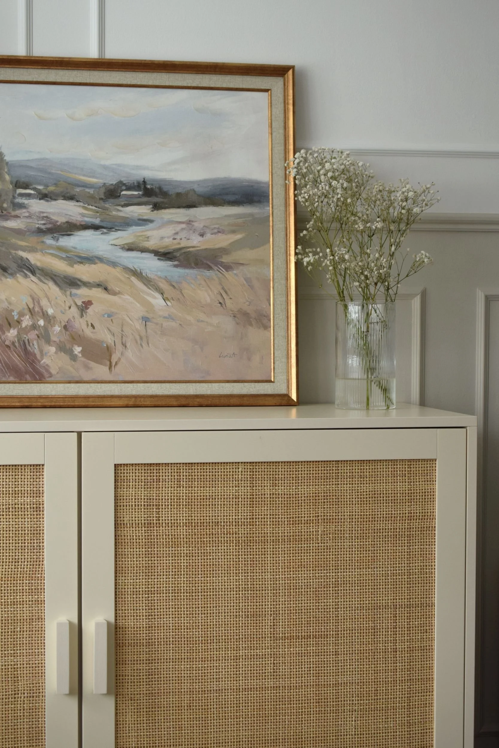 A painting of a landscape with a river, hills, trees, and cloudy sky, framed in wood, is hung on a white wall. To the right, a vase with white baby's breath flowers sits on a white dresser with dark rattan panels.