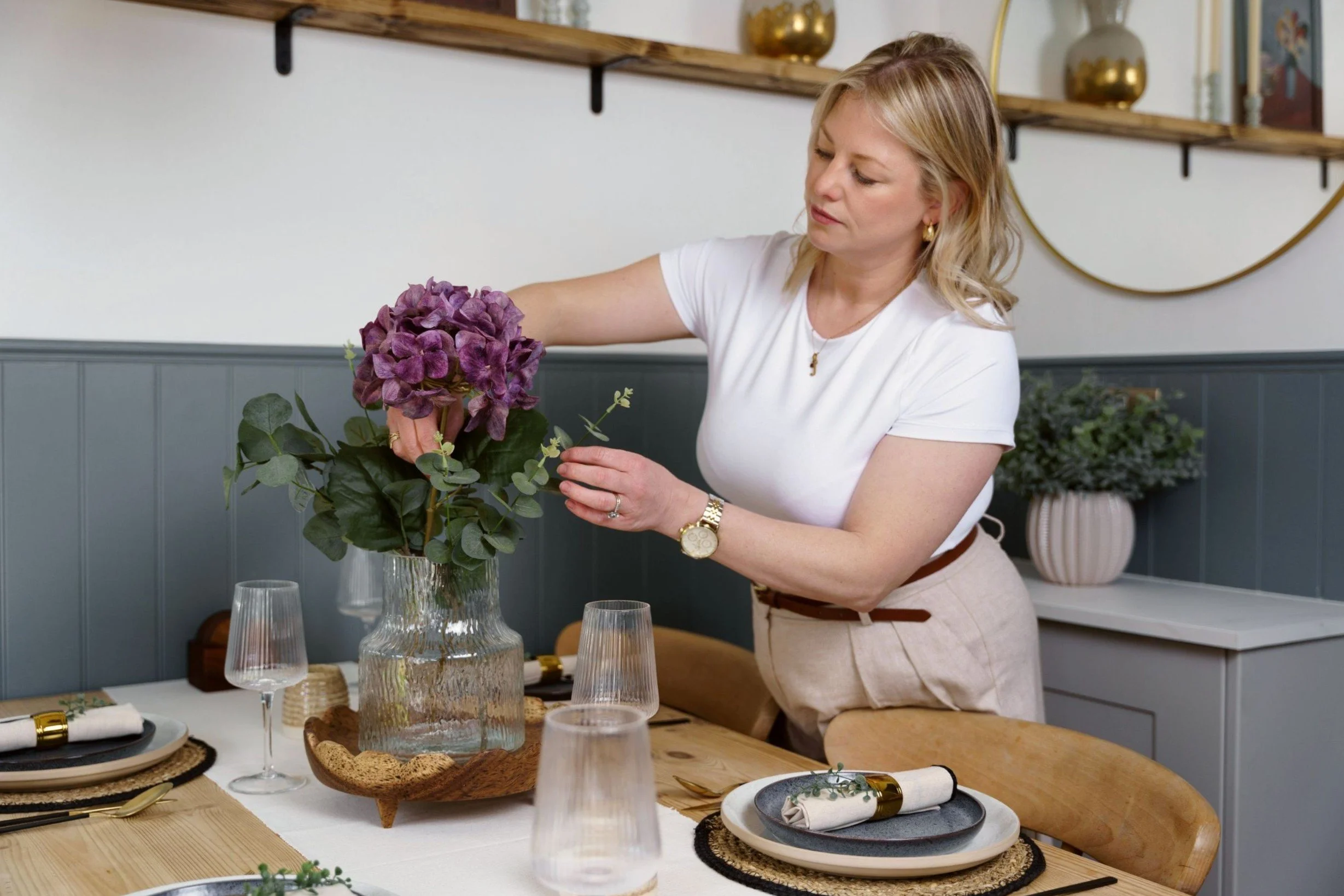 A woman with blonde hair, wearing a white t-shirt and beige skirt, is arranging a purple hydrangea flower in a glass vase at a dining table set for a meal, with place settings and wine glasses in a modern, decorated room.