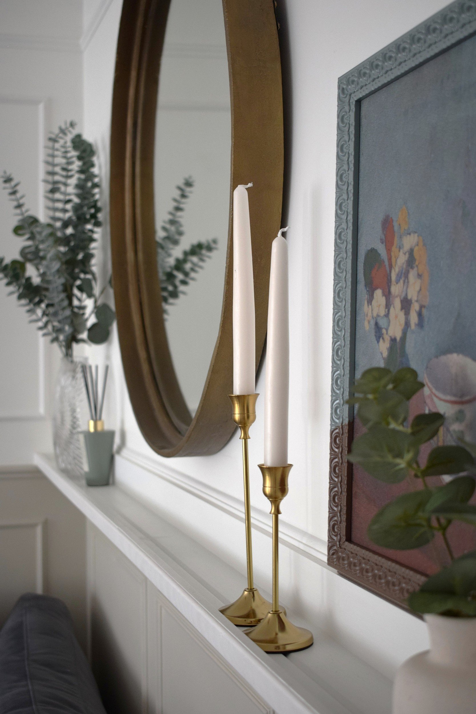 A mirror on a wall with a gold frame, reflecting a white wall with decorative molding and some plants in vases, including eucalyptus, on a white fireplace mantel.