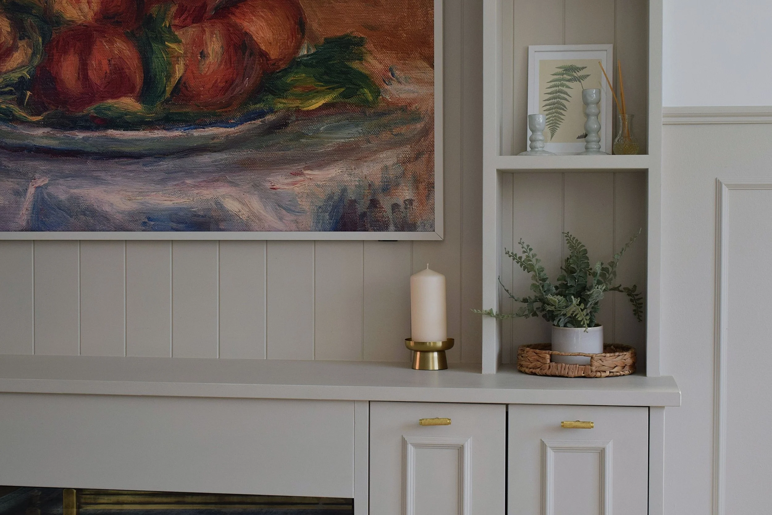 A white wall cabinet with a shelf on top, decorated with a framed botanical print, two white candles in candlesticks, a glass vase with reed diffuser sticks, and a potted plant with green and gray foliage. Part of a large, colorful still life painting is visible on the left side of the image.