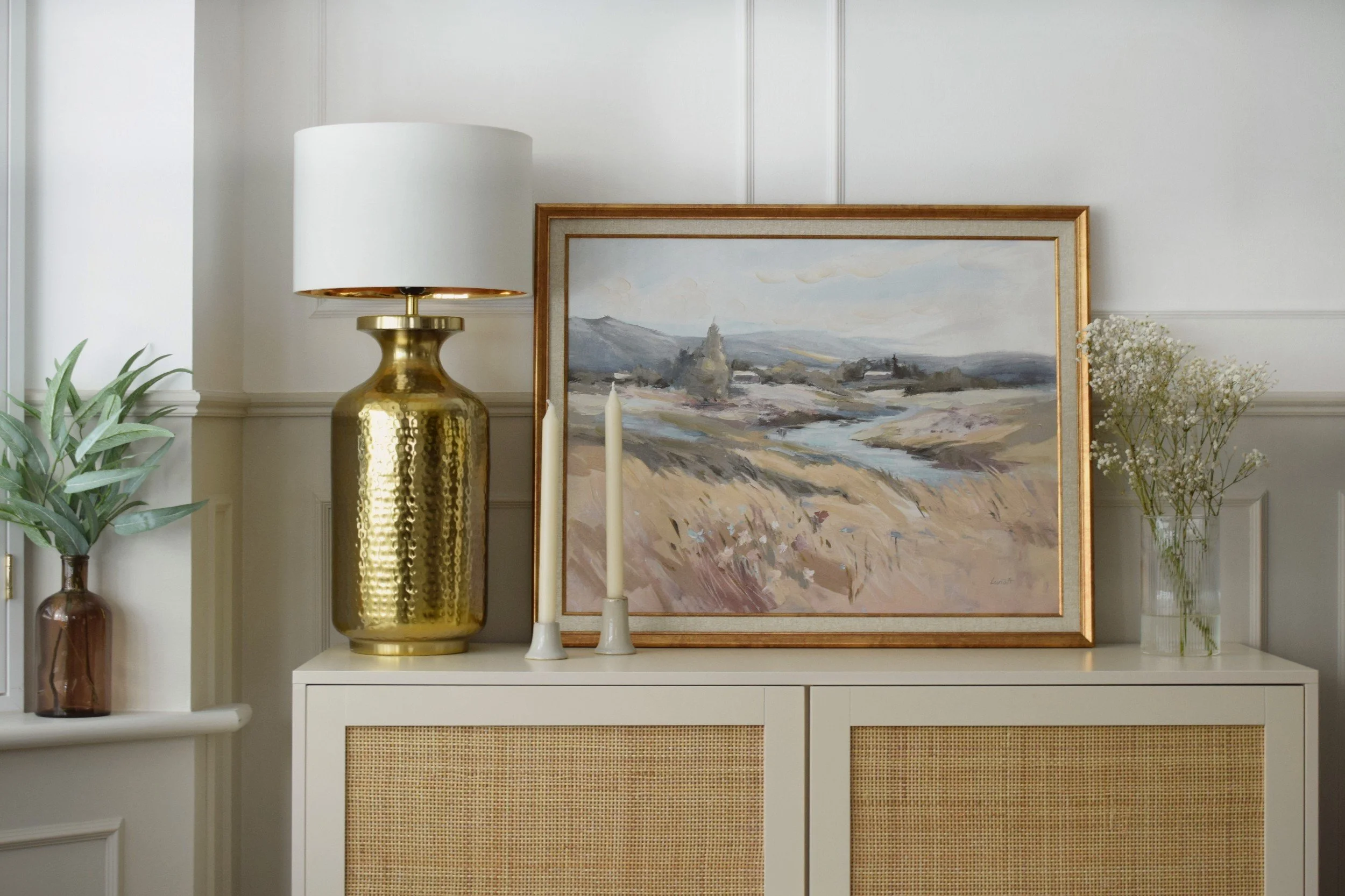 Decorative living room corner with a white cabinet, a large gold vase, a painting of a rural landscape, two white candles, a large white lampshade, green plants in a brown vase, and a bunch of white flowers.