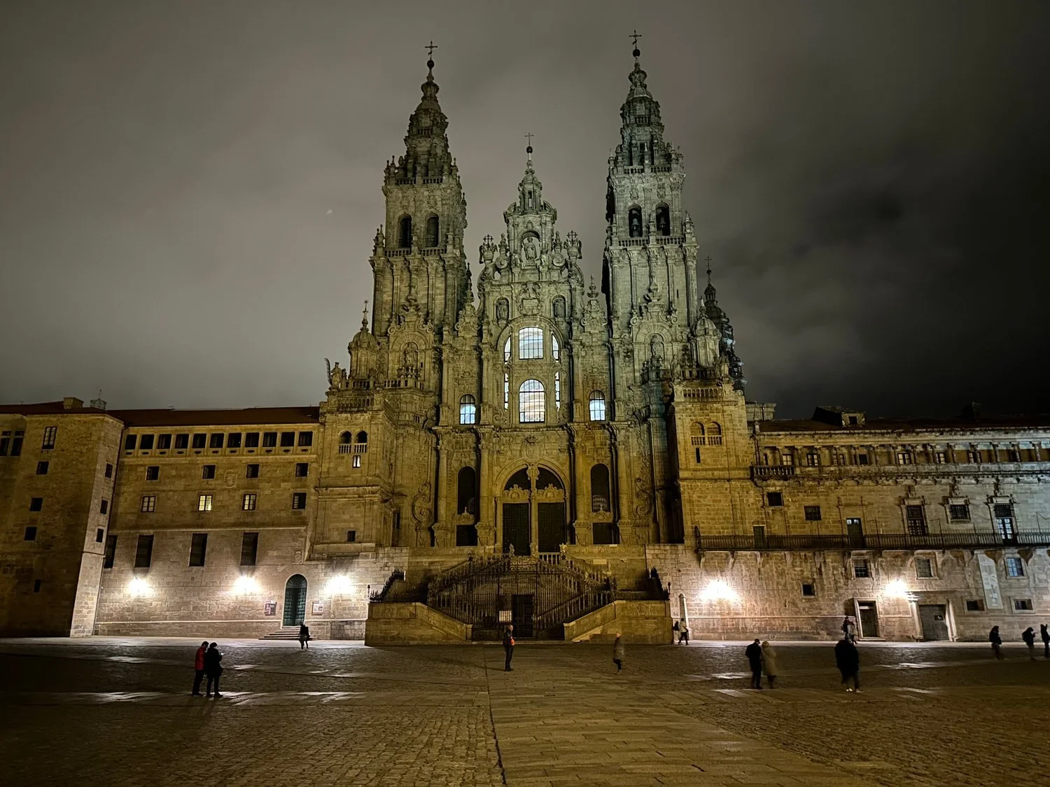 The Obradoiro facade of Santiago de Compostela Cathedral lit at night, its twin towers and double staircase rising over a nearly empty stone square.