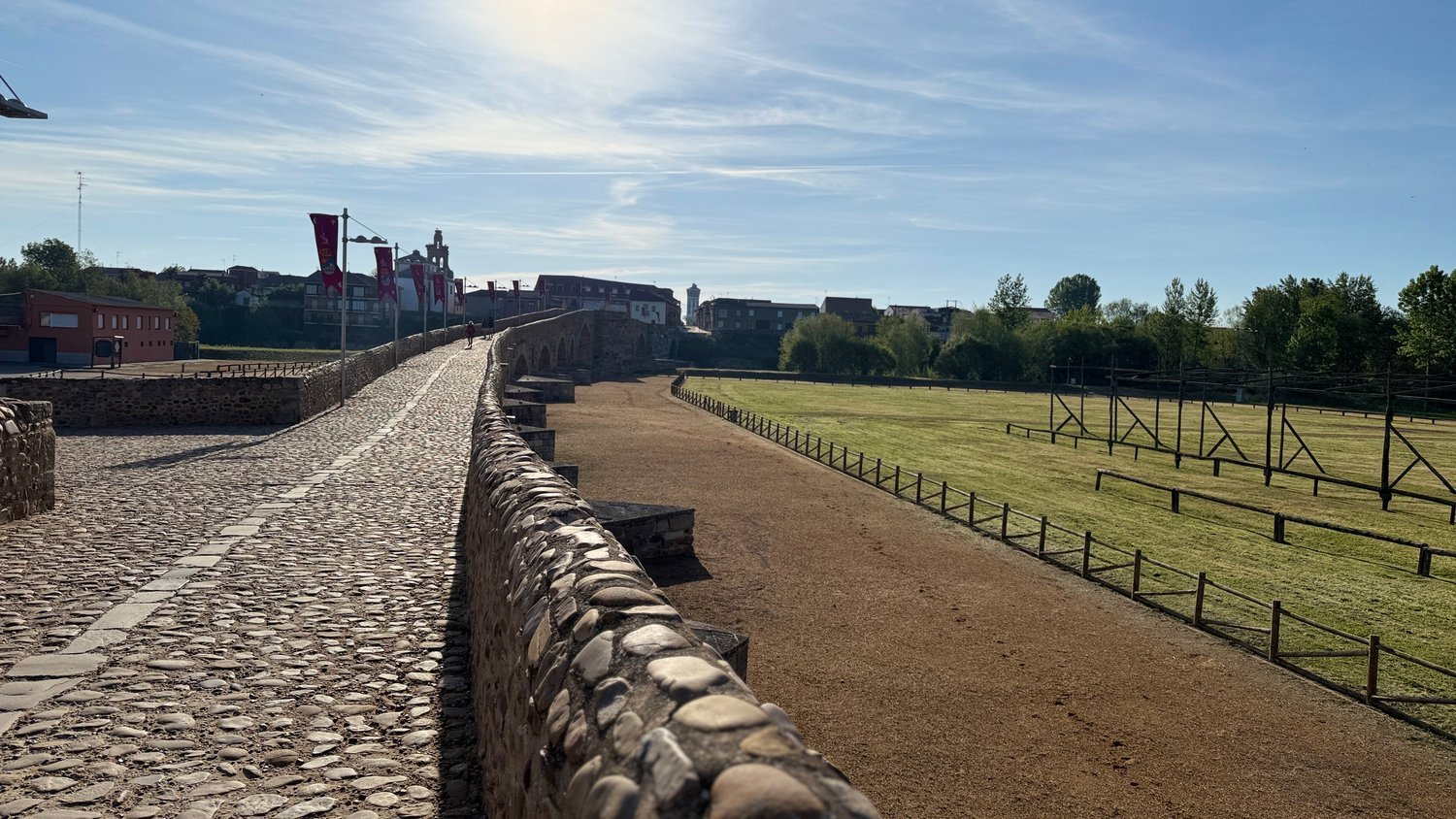 A long cobbled stone bridge in morning sun, red banners along its side, with a grass field and wooden fencing below.