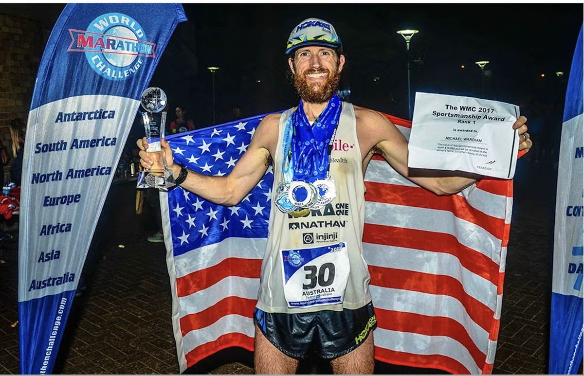 Ultrarunner Michael Wardian draped in an American flag, holding a trophy and Sportsmanship Award certificate, standing between World Marathon Challenge continent banners in Sydney, Australia.