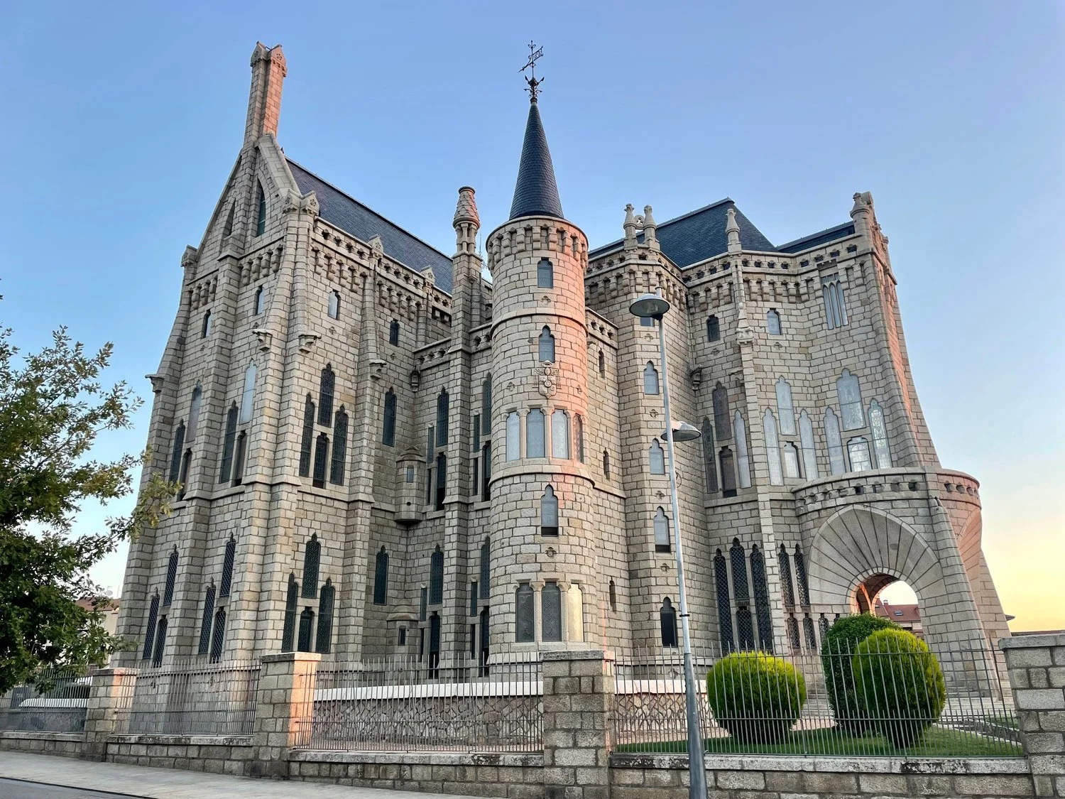 Gaudí's Episcopal Palace in Astorga, a gray stone building with a pointed slate turret, tall arched windows and a stone archway, behind an iron fence in early morning light.