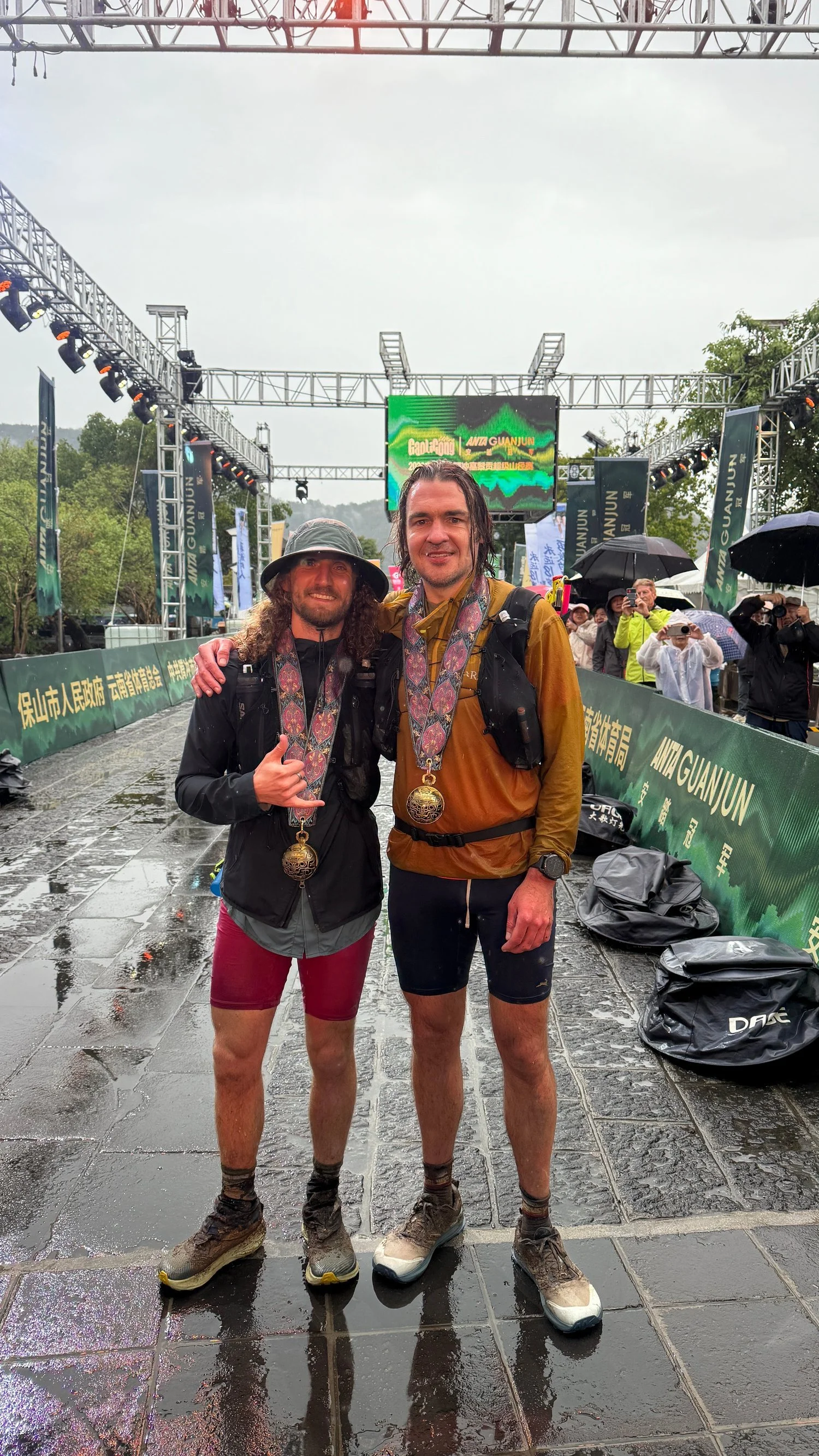Gabriel Green and Matt Gaide standing in the rain at the Mt. Gaoligong Ultra finish line wearing finisher medals.