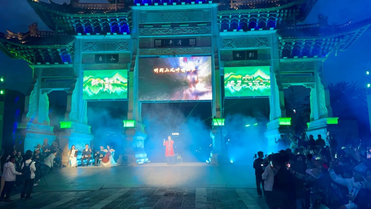 Lit stone archway in Tengchong at night with a singer, string quartet, video screens and stage smoke during the Mt. Gaoligong Ultra opening ceremony.