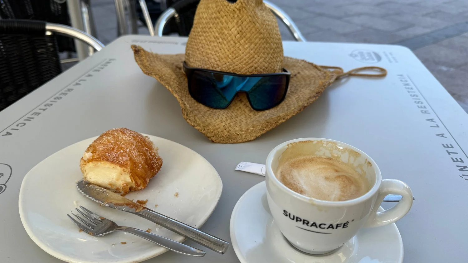 A half eaten pastry with a sugared crust and cream filling on a plate beside a nearly finished coffee, with a straw hat and sunglasses on a cafe table.
