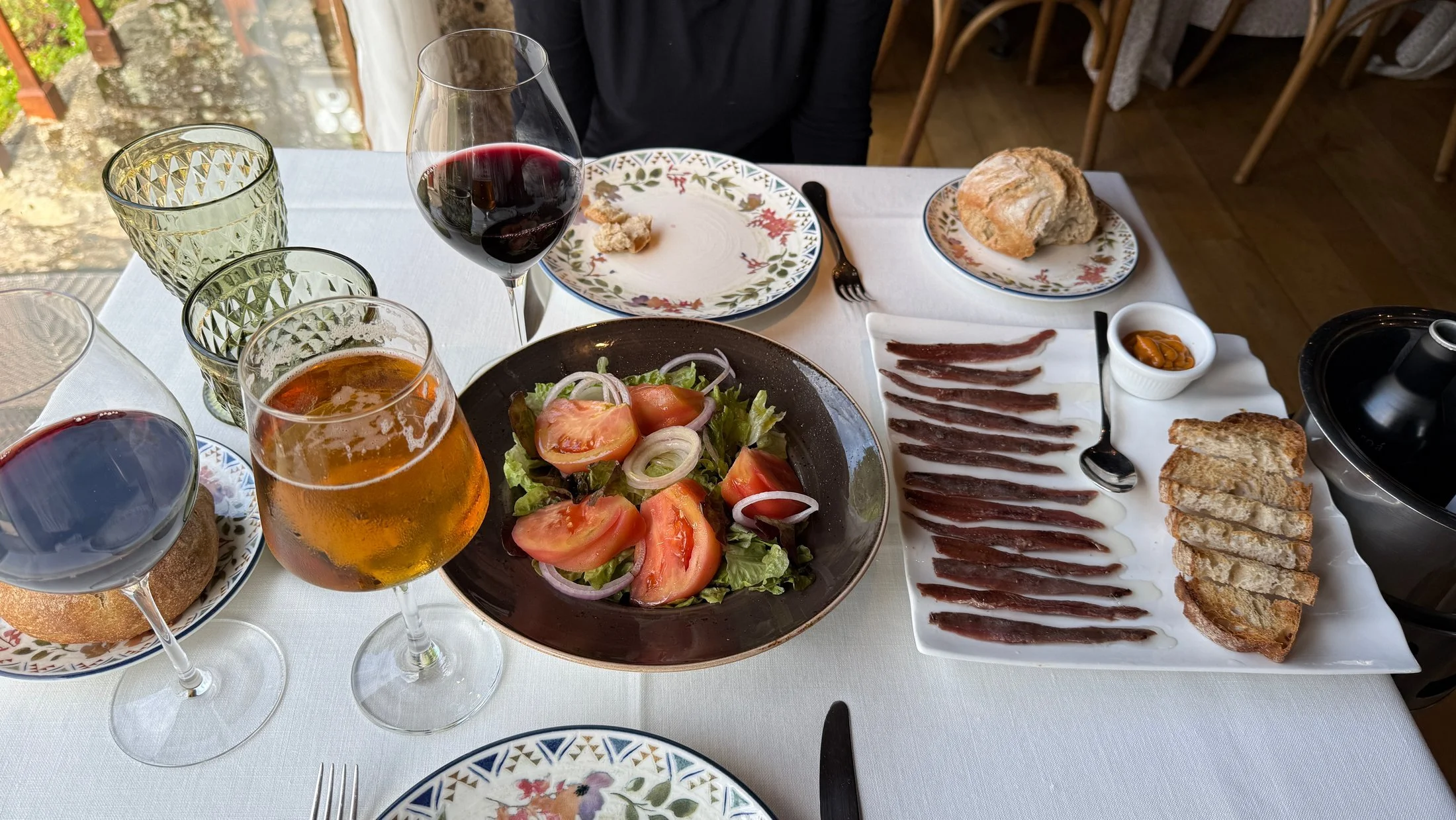 A table at Fervenza Casa Grande in O Corgo set with a salad of tomato, onion and lettuce, a plate of anchovy fillets with toasted bread, a bread roll, two glasses of red wine and a glass of amber beer.