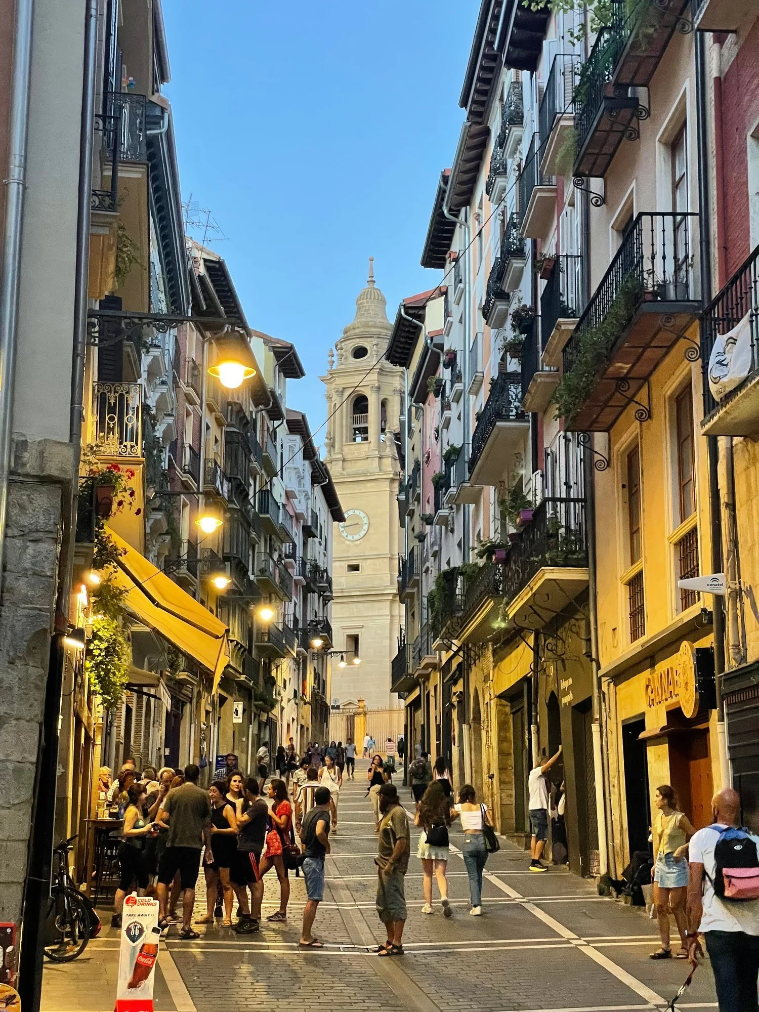 A narrow street in Pamplona's old town lined with balconied buildings and lit lamps, people gathered outside a bar and a stone bell tower with a clock at the far end.