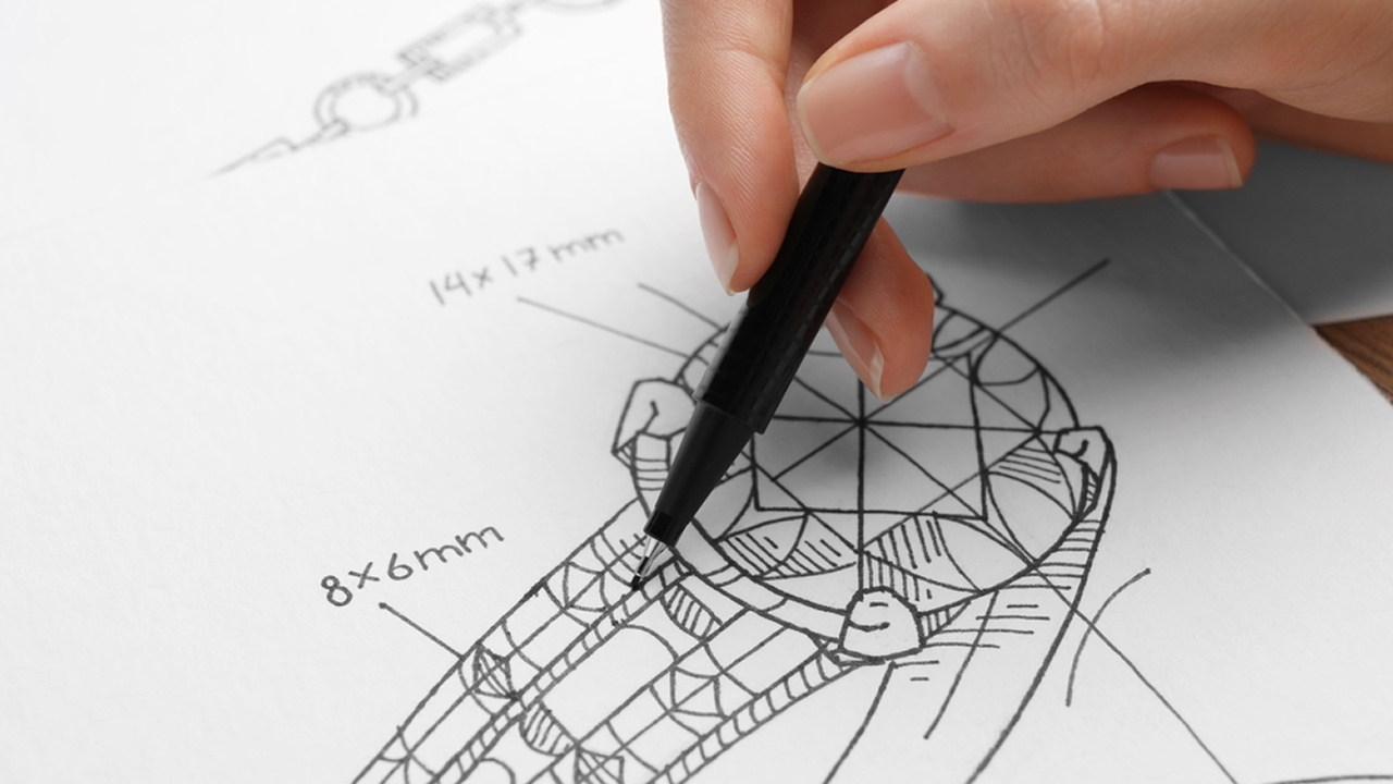 Jewelry designer hand-drawing a detailed custom engagement ring sketch with gemstone millimeter measurements.