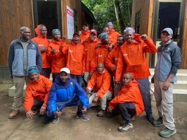 Group of people wearing orange jackets posing outdoors in front of a wooden building.