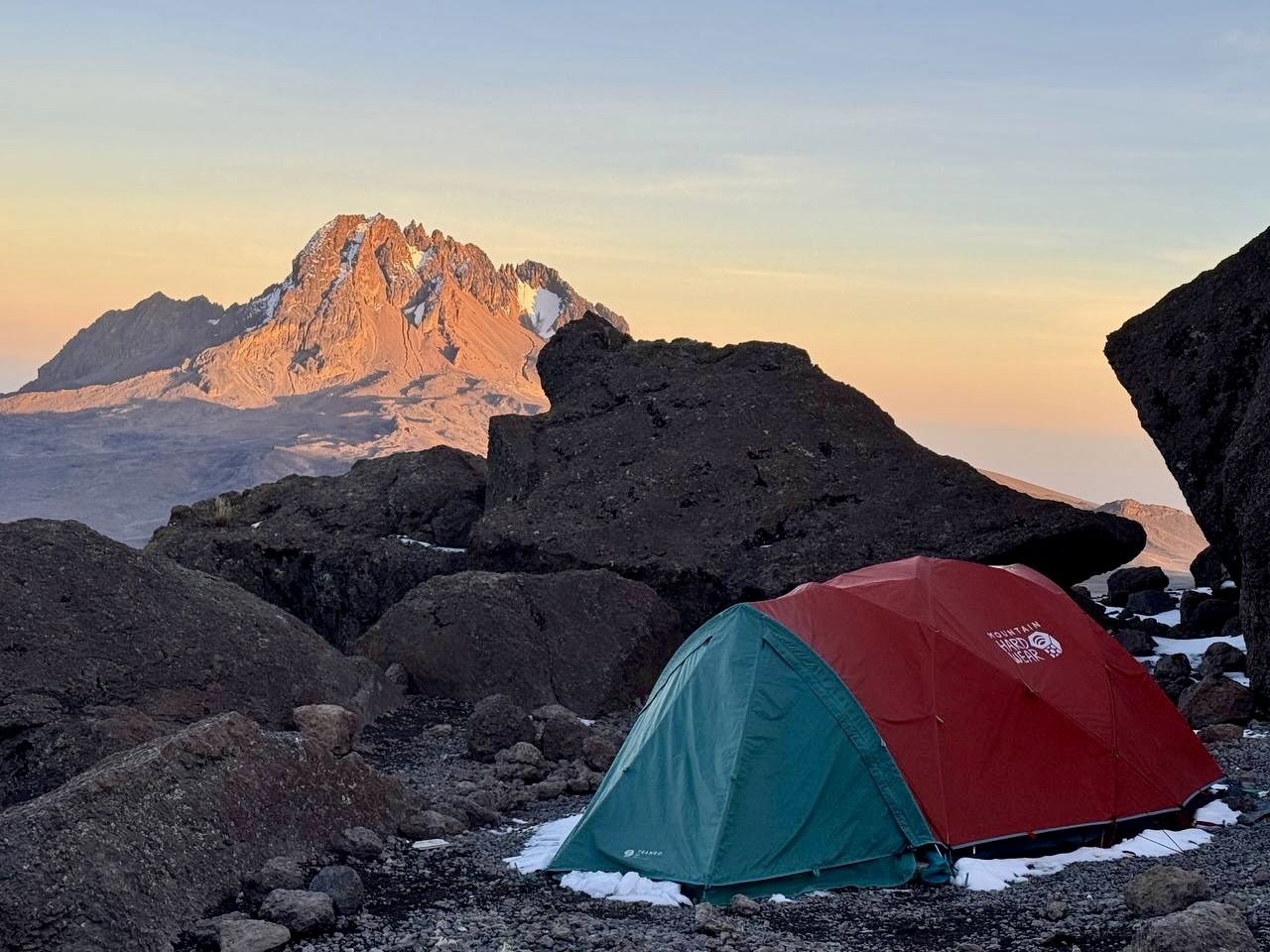 A red and blue tent set up on rocky terrain surrounded by large boulders with a mountain in the background during sunset.