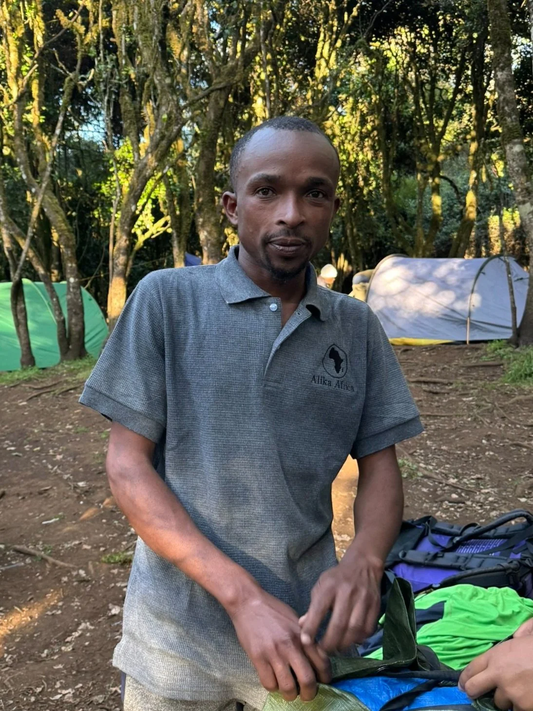 Person in gray shirt outdoors setting up camping gear with tents and trees in the background.