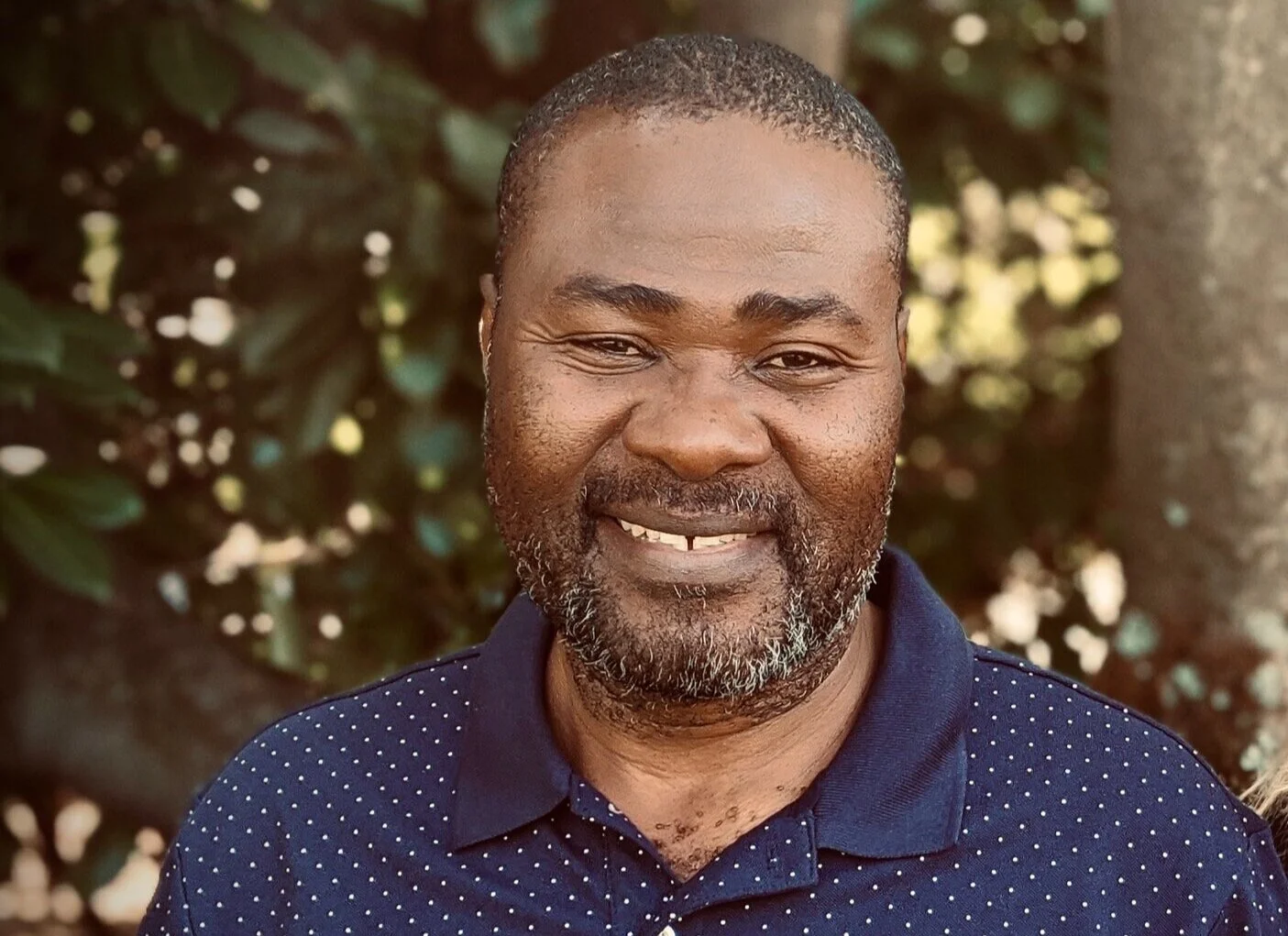 Smiling man wearing a blue polka-dotted shirt standing outdoors with trees in the background.