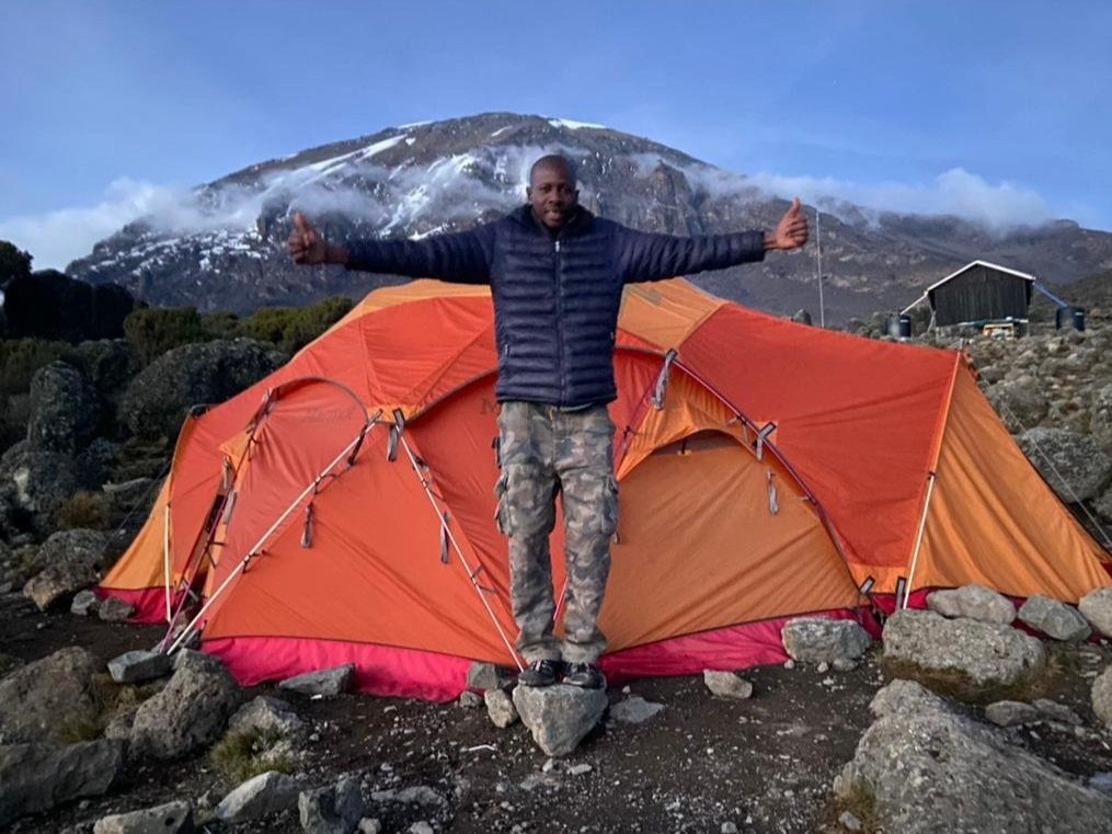 Man standing in front of orange tents on rocky terrain with a mountain in the background, arms extended, wearing a dark jacket and camouflage pants.
