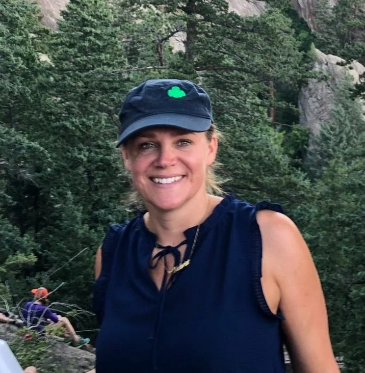 Smiling person wearing a black cap and navy sleeveless top standing in front of a forested mountain landscape.